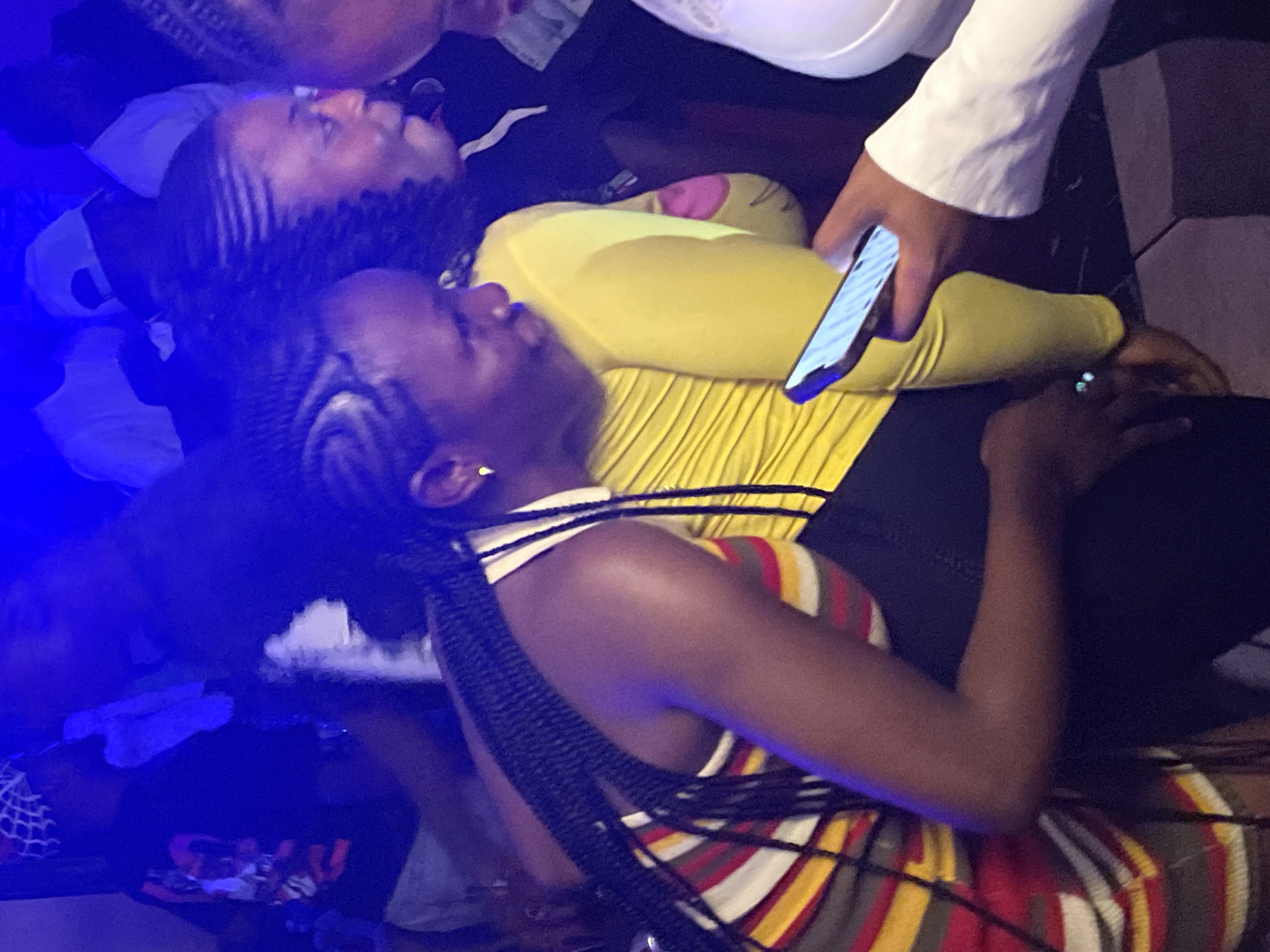 A group of young African adults are captured in a dimly lit, indoor setting, possibly a nightclub or party, in Durumi, Nigeria. The blue ambient lighting casts a vibrant glow over the scene. In the foreground, a woman with braided hair wears a multicolored striped dress and a black skirt. She is holding a smartphone, its screen illuminated, and her attention is directed towards it. To her right, another woman in a bright yellow top is partially visible, her face turned away from the camera. Behind them, other individuals are faintly discernible, some wearing white shirts and caps. The overall impression is one of a lively social gathering, with a focus on technology and personal interaction within a vibrant urban nightlife environment.