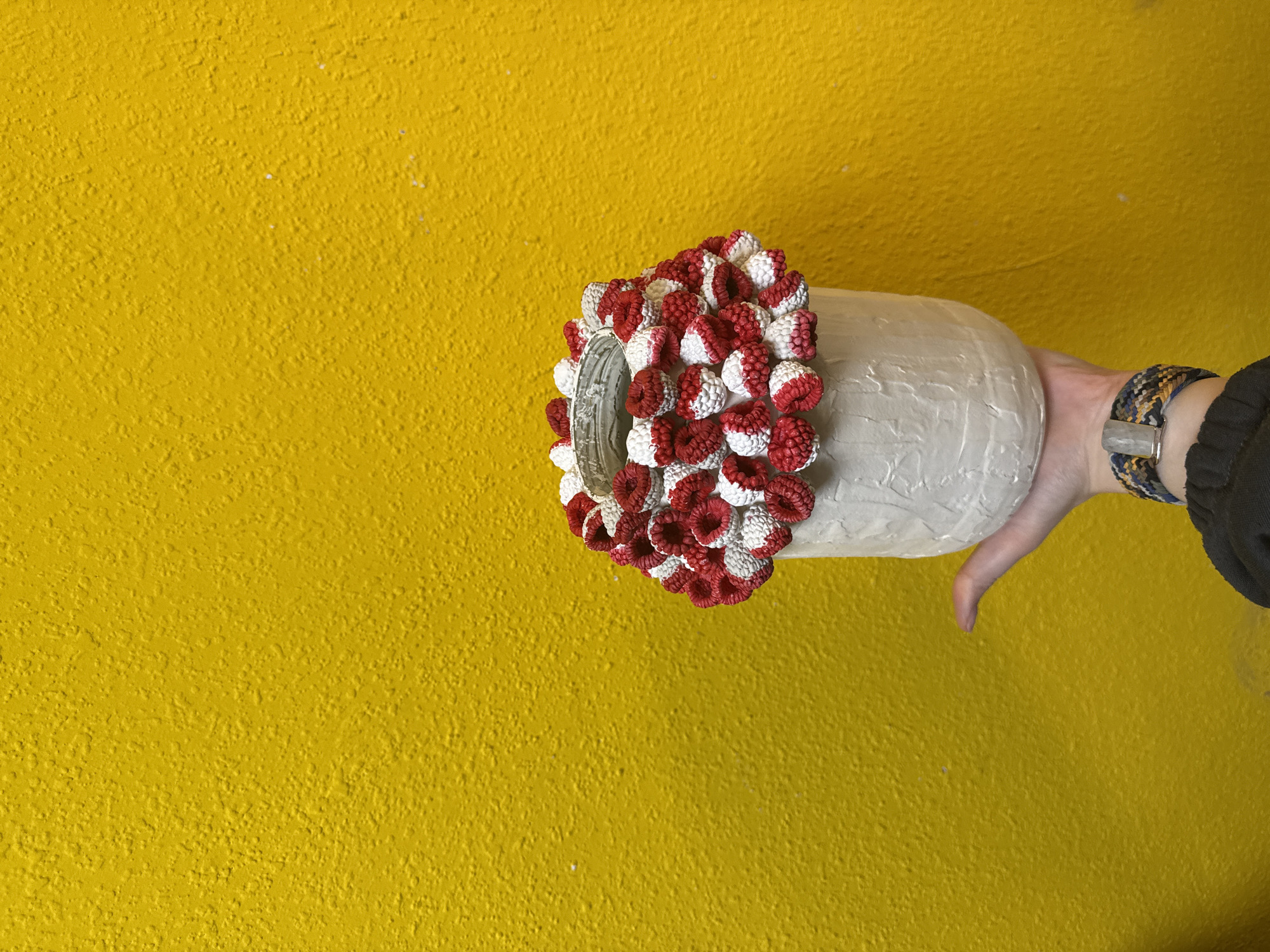 A hand holds a textured white vase adorned with red and white decorative elements against a yellow textured wall. The hand wears a multi-colored woven bracelet with a silver clasp. The location is not specified and therefore Unknown, Unknown.