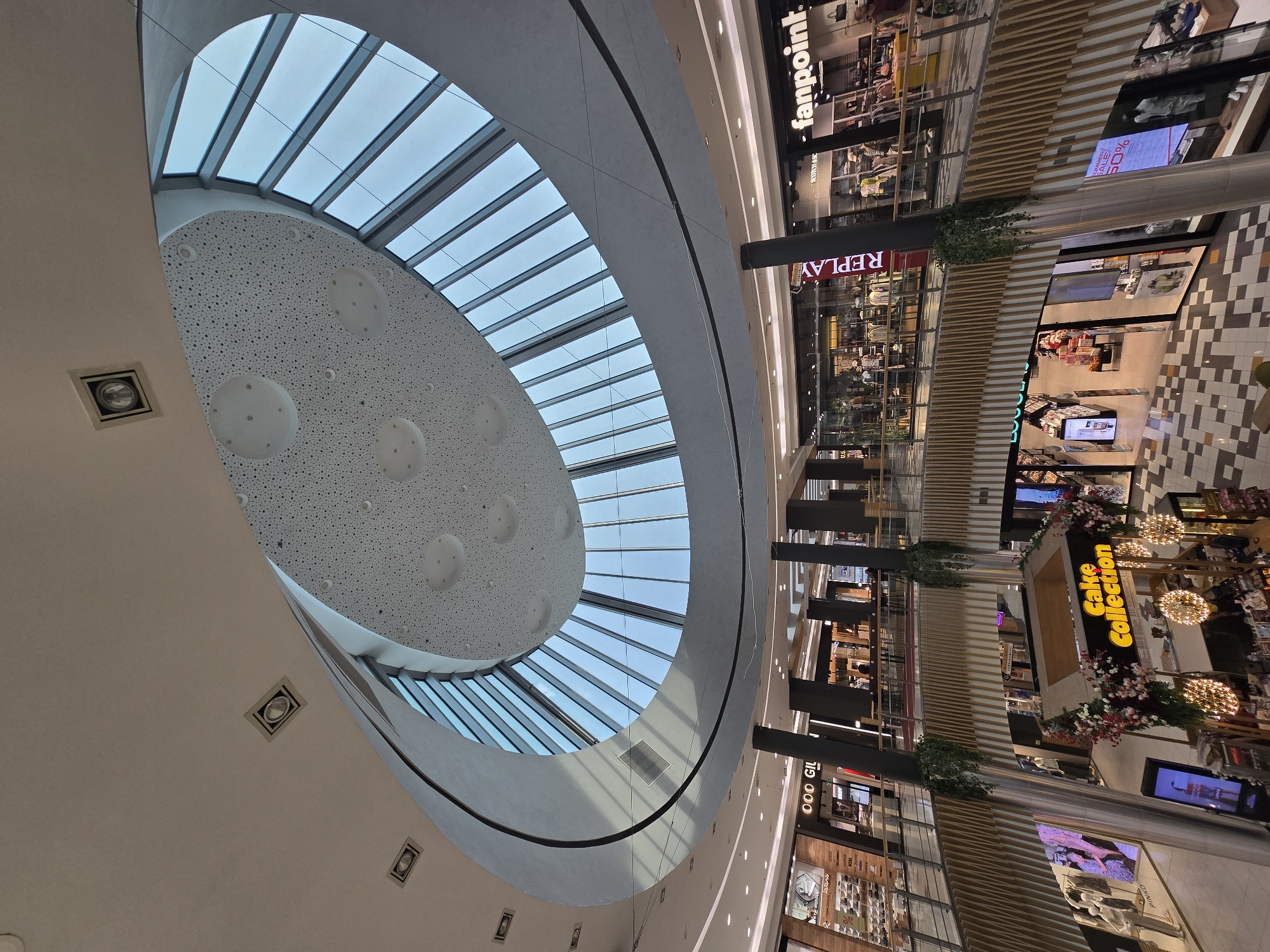 This image depicts the interior of a multi-level shopping mall, likely located in Plovdiv, Bulgaria, characterized by its modern architecture and extensive use of glass.
The central feature is a large, circular atrium with a domed, sloped glass ceiling, divided into many paneled sections, allowing natural light to flood the space. Below this, suspended structures and walkways connect different levels of the mall.
Several shops are visible on multiple floors, with prominent signage such as "fanpoint" and "REPLAY" indicating retail outlets. The shops display clothing, accessories, and other merchandise, with mannequins visible in some storefronts.
The lower levels show the mall's flooring, a patterned checkered design. Decorative elements, including hanging plants and floral arrangements around a stall labeled "Cake Collection," add to the ambiance.
Lighting fixtures are integrated into the ceiling and walkways, illuminating the interior. Some ceiling areas feature perforated panels with embedded lights.
People are present within the mall, browsing shops and walking on the walkways, though they are not the primary focus of the image. The overall impression is one of a contemporary and bustling commercial environment.