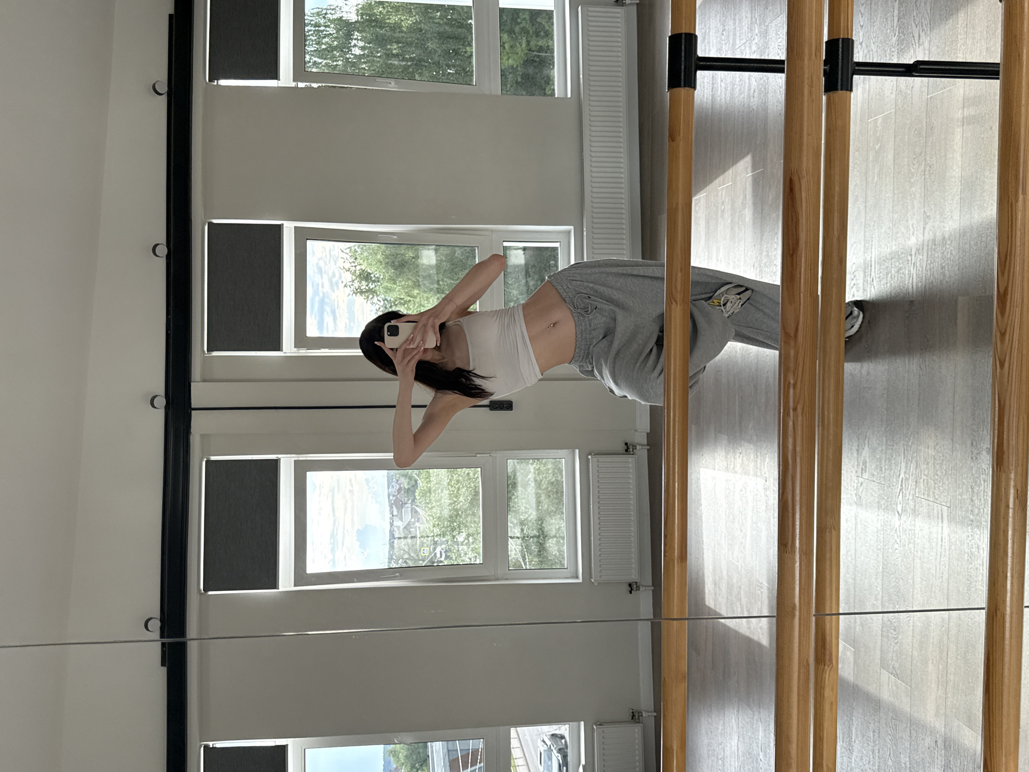 This media file shows a full-body mirror reflection of a young woman in a dance studio. She is wearing a white cropped tank top and grey sweatpants, with a small piercing visible on her navel. She is holding a smartphone, taking a selfie, and her expression appears neutral or slightly playful. The room features large windows that offer a view of trees and greenery, suggesting it's daytime and likely fair weather. The studio has a polished wooden floor and a barre along one wall, indicated by the vertical wooden poles. The presence of radiators under the windows and the overall aesthetic suggest a modern, clean space designed for physical activity. The reflection also shows part of a black metal bar attached to the wooden poles, part of the barre structure.