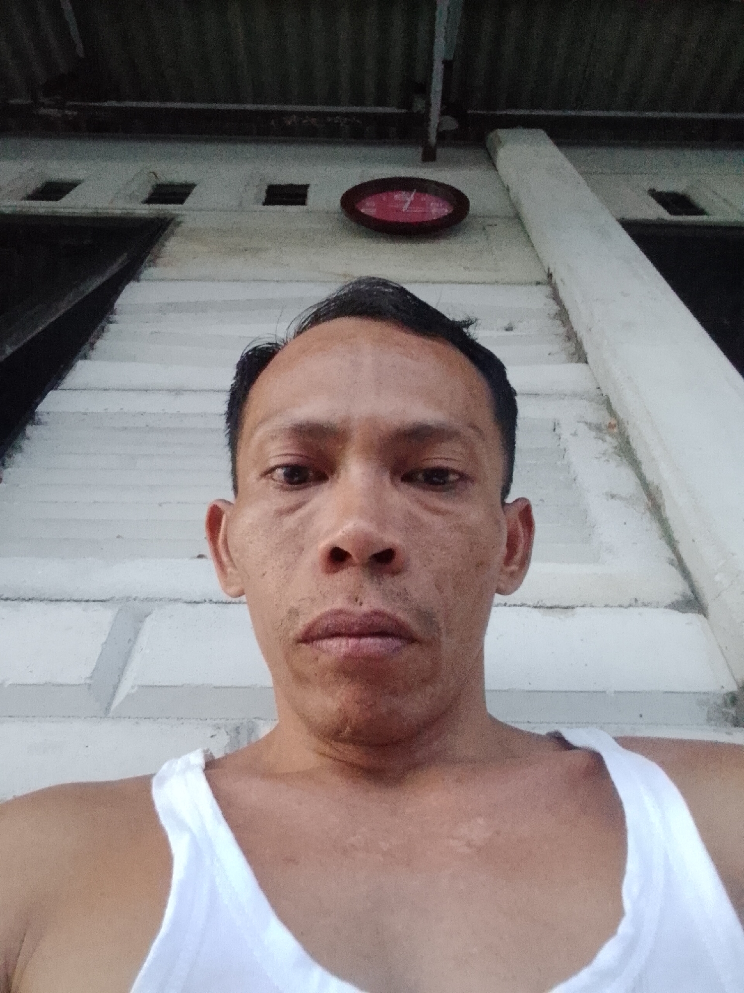 A middle-aged man in a white undershirt is taking a selfie. He has short dark hair and light brown skin. Behind him is a white wall with small square windows and a red clock. The roof appears to be corrugated metal. The location is Bukit Ketok, Indonesia.