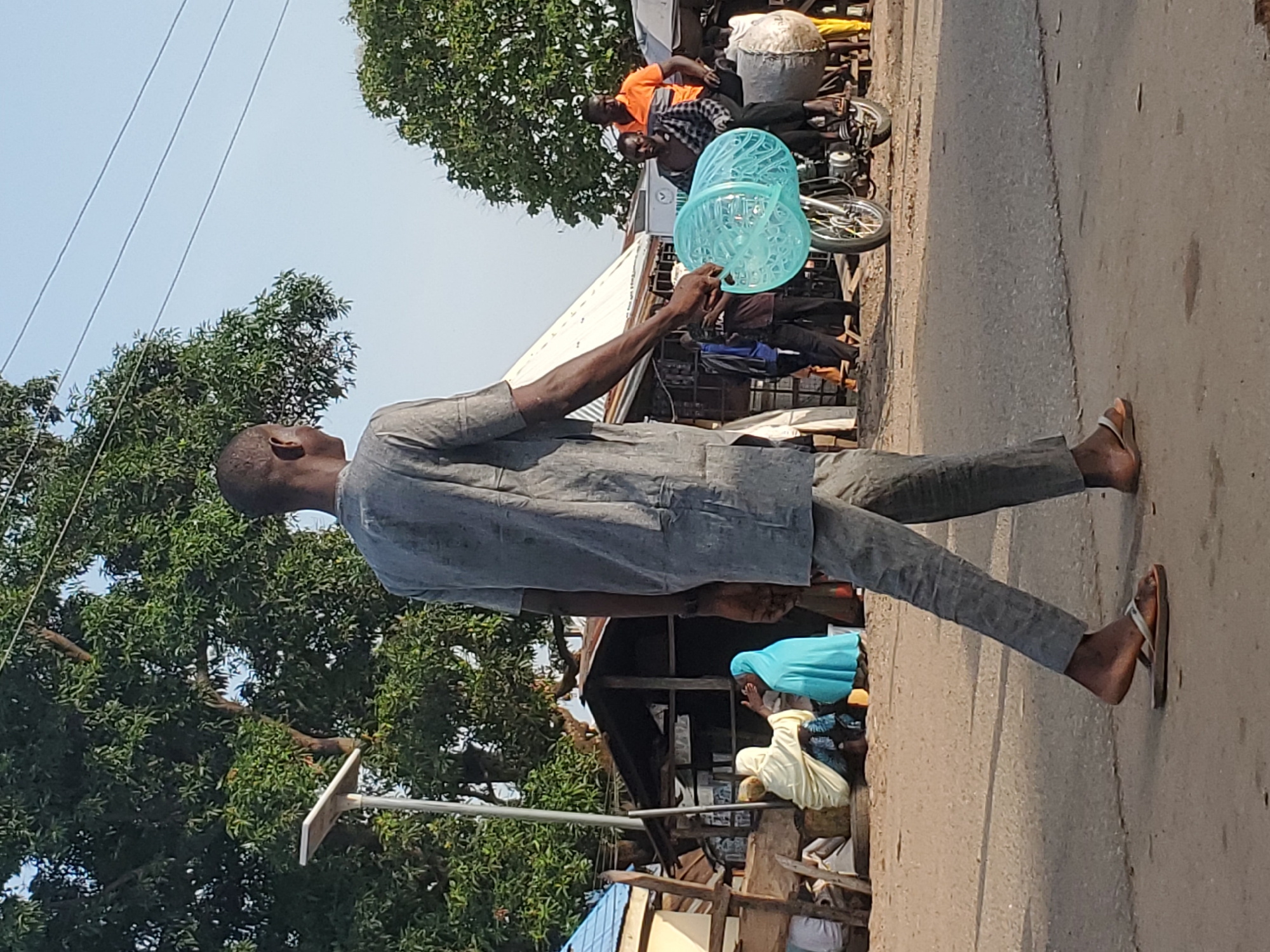 In Mararaba, Nigeria, a man walks along a paved road carrying a stack of light blue plastic bowls. He is dressed in gray clothing and wears flip-flops. Behind him, a motorcycle carries cargo and two individuals, one wearing an orange shirt. Nearby, two women are visible in a partially obscured structure. Trees line the roadside under a clear sky.
