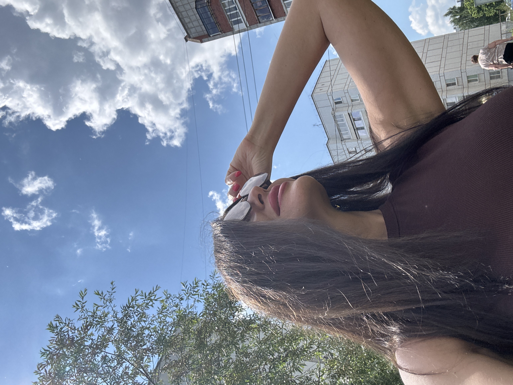 An outdoor, low-angle, medium shot captures a woman with long, dark hair wearing a brown tank top and sunglasses. She is holding her right hand up to her sunglasses, shielding her eyes from the sun. The background shows a clear blue sky with white clouds, a tree on the left, and apartment buildings on the right. The scene appears to be set in a residential area of Chaikovsky, Russia, given the architecture of the buildings.