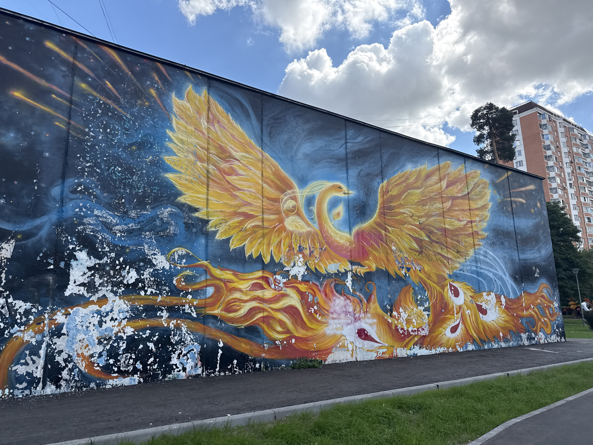 The image shows a mural painted on the side of a building in Lyubertsy, Russia. The mural depicts a large, golden phoenix rising from flames against a dark blue, starry background. The phoenix has its wings spread wide, and its feathers are rendered in shades of yellow and orange. Below the phoenix, flames are depicted in vibrant reds and oranges, with some white and pink highlights. The lower portion of the mural shows significant peeling and damage to the paint, revealing the concrete beneath. In the background, to the right, a tall, multi-story apartment building is visible, along with trees and a lamppost. The sky is blue with scattered white clouds. A paved walkway and a strip of green grass are in the foreground.