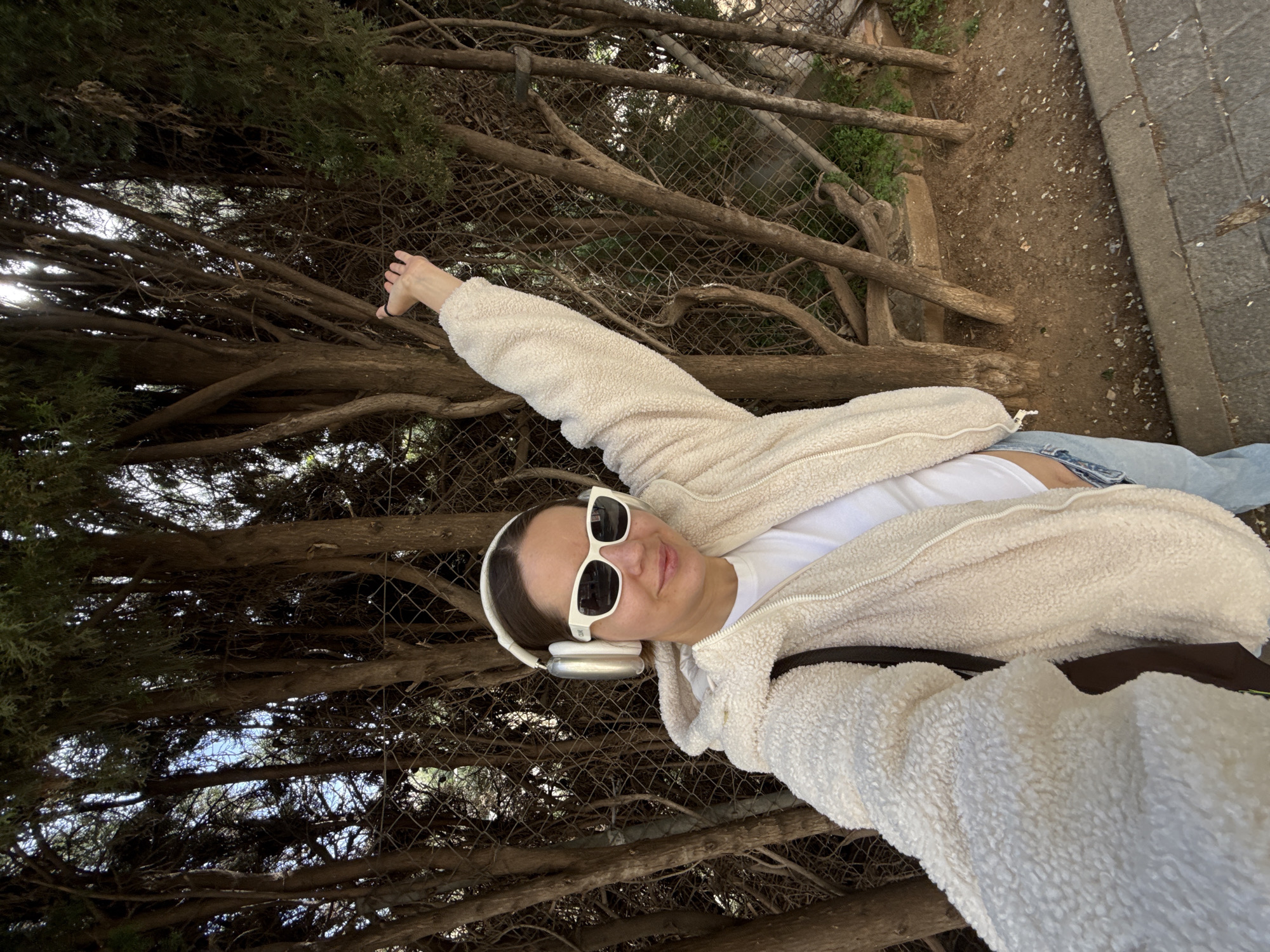 A medium-shot, rotated selfie of a fair-skinned person with light brown hair, wearing a white fluffy zip-up jacket, a white t-shirt, blue jeans, white sunglasses, and white headphones. The person's left arm is extended and raised towards the top-left corner of the frame. The background includes an area with many branches, small trees, and a metal chain-link fence. To the right of the frame, there is brown dirt and a grey stone structure. The image was possibly taken in or near the city of Barcelona, Spain.