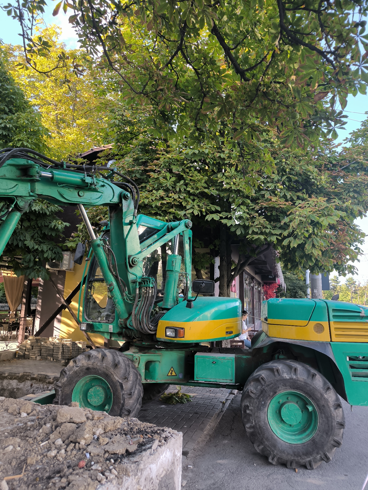 The image shows a green and yellow excavator parked on a paved sidewalk next to a street. There are piles of dirt and construction materials nearby. Trees with green and yellow leaves hang over the scene, and a building with windows and a yellow awning is visible in the background. A person is seated at a table outside the building. The excavator is the primary subject of the image, and it appears to be in a working or construction zone. The overall scene suggests an urban environment undergoing some form of renovation or repair. This photo was taken in Chișinău, Moldova.