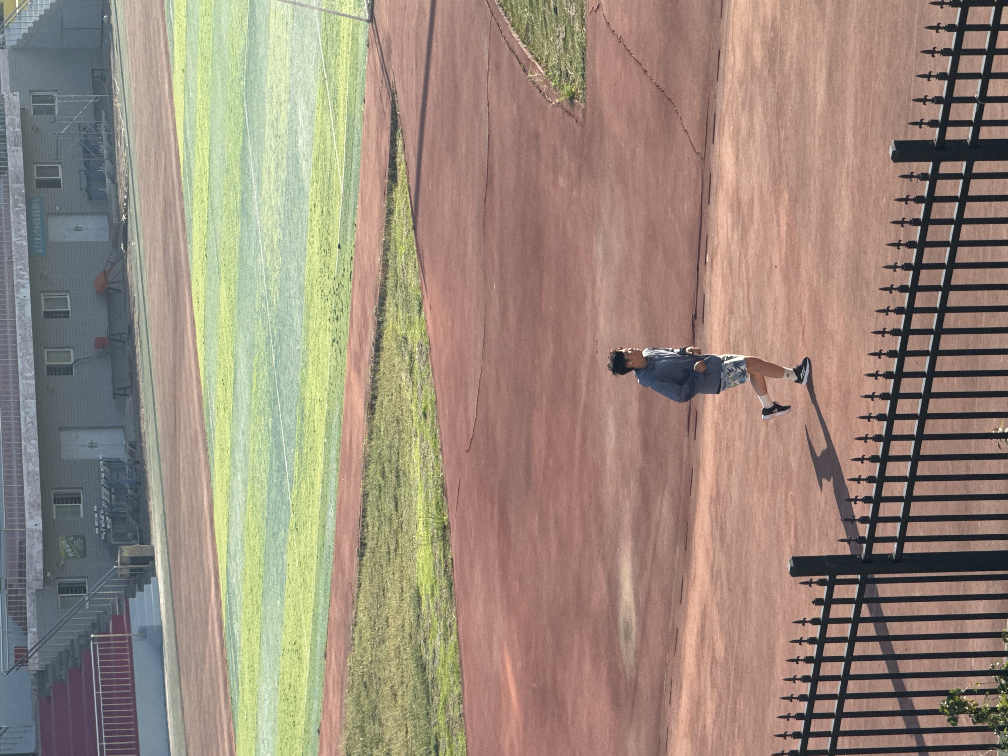 The image depicts an outdoor track and field facility, likely located in Zhongtun, China, as indicated by the presence of Chinese text on the building in the background. A young person, dressed in athletic wear, is actively jogging on the reddish-brown track in the foreground. The person is wearing a light blue hooded jacket and patterned shorts. The track is bordered by a black metal fence in the lower portion of the image. Behind the track, a section of green turf, possibly a sports field, is visible, interspersed with red boundaries. In the distance, a building with multiple windows and doorways stands, along with stadium seating on the left side. The lighting suggests it is either early morning or late afternoon, casting a shadow of the jogger on the track. The environment appears to be a sports complex designed for athletic training and competition.