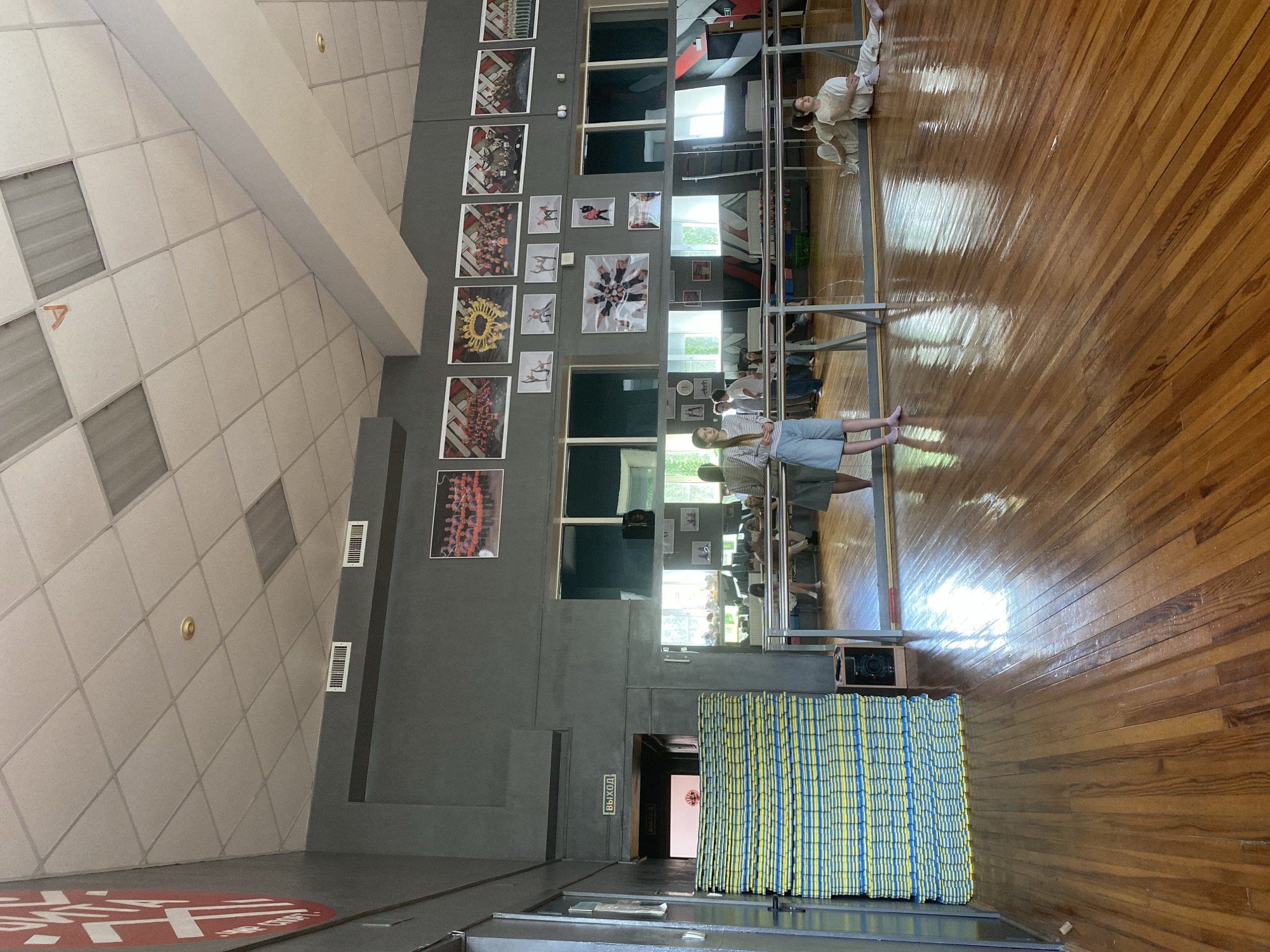 The image depicts the interior of a dance or martial arts studio. The room features a polished wooden floor, large mirrors along one wall, and a high ceiling with acoustic tiles and lighting fixtures. Several framed photographs of individuals in various poses are displayed on the wall. In the foreground, a stack of blue and yellow mats is visible. Three young women are present in the scene; two are standing near the mirrors, and one is sitting on the floor. One of the standing women is wearing a striped long-sleeved shirt and denim shorts, while the other is in a white t-shirt and denim shorts. The seated woman is in white athletic wear. The studio appears to be in the city of Votkinsk, Russia, as suggested by the "ВЫХОД" (Exit) sign above a doorway.
