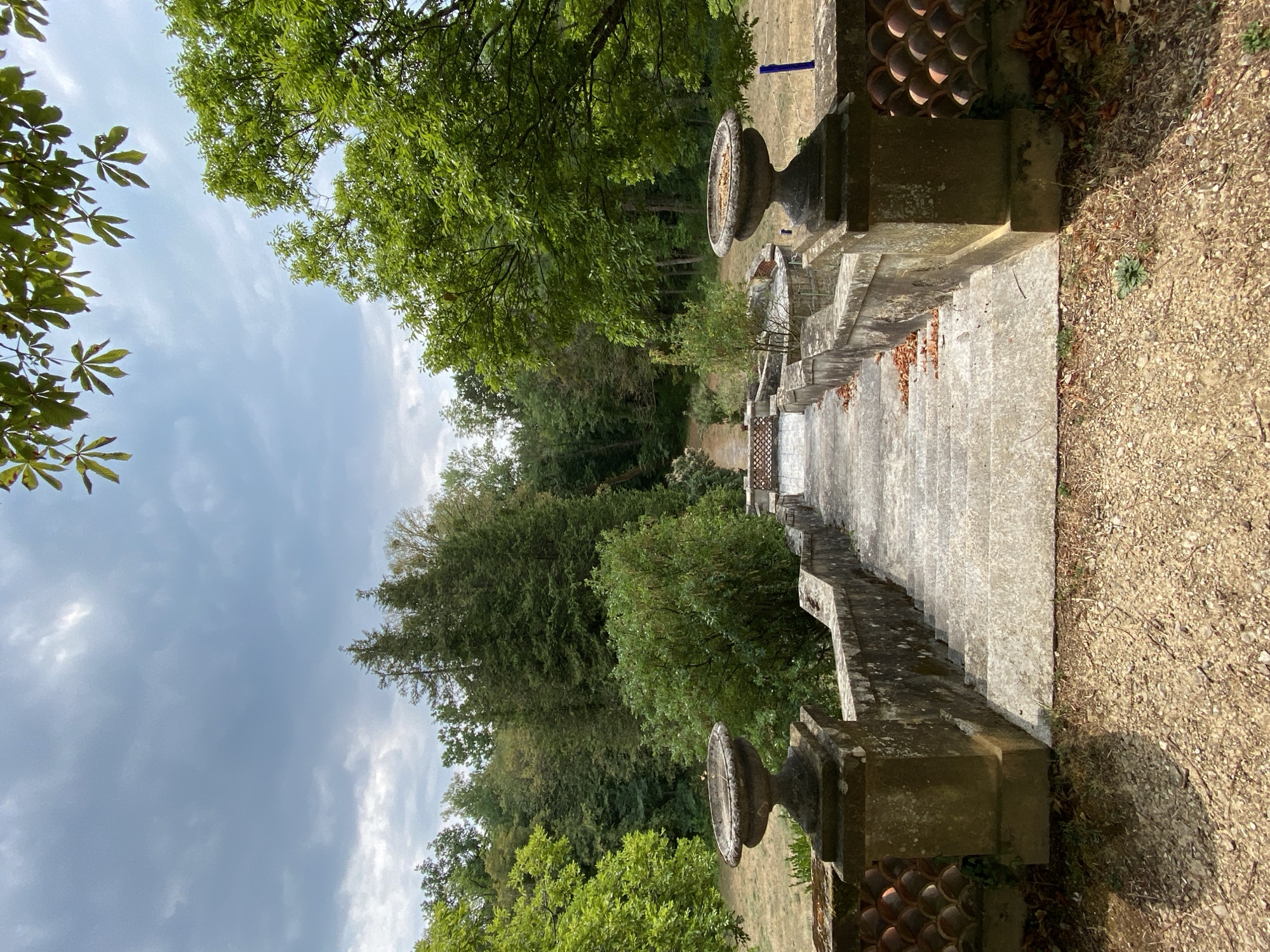 The image depicts an outdoor view of a stone staircase leading down into a wooded area. The staircase is flanked by stone balustrades with decorative urns at the top. The balustrades are partially covered in moss and have a decorative terracotta tiling pattern along the lower section. The ground leading up to the stairs is covered in gravel and scattered dry leaves. Beyond the staircase, the path continues into a forest of various trees, with a patch of dry, mowed grass to the right. The sky above is cloudy and overcast. The scene appears to be a garden or park setting, possibly at the historical site of Laténa in Switzerland, given the context. There are no people or animals visible in the image.