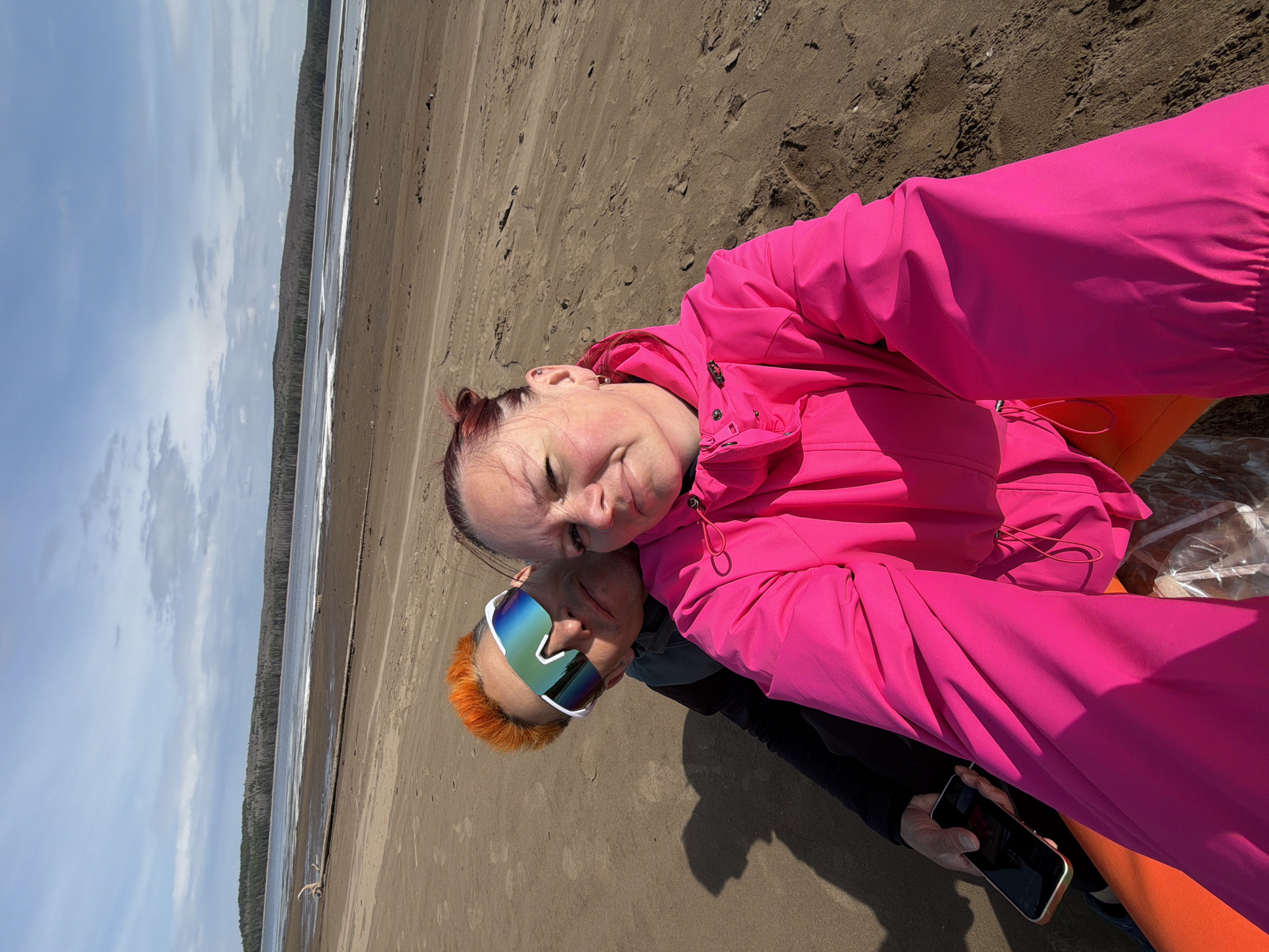 A photograph shows two individuals on a beach. The person in the foreground is wearing a bright pink jacket. The other person is wearing mirrored sunglasses and has orange hair. They appear to be taking a selfie with a smartphone, which is visible in the lower right corner of the image. The background shows a beach with a body of water alongside a treeline. It is known that this is in the city of Chaikovsky, Russia.