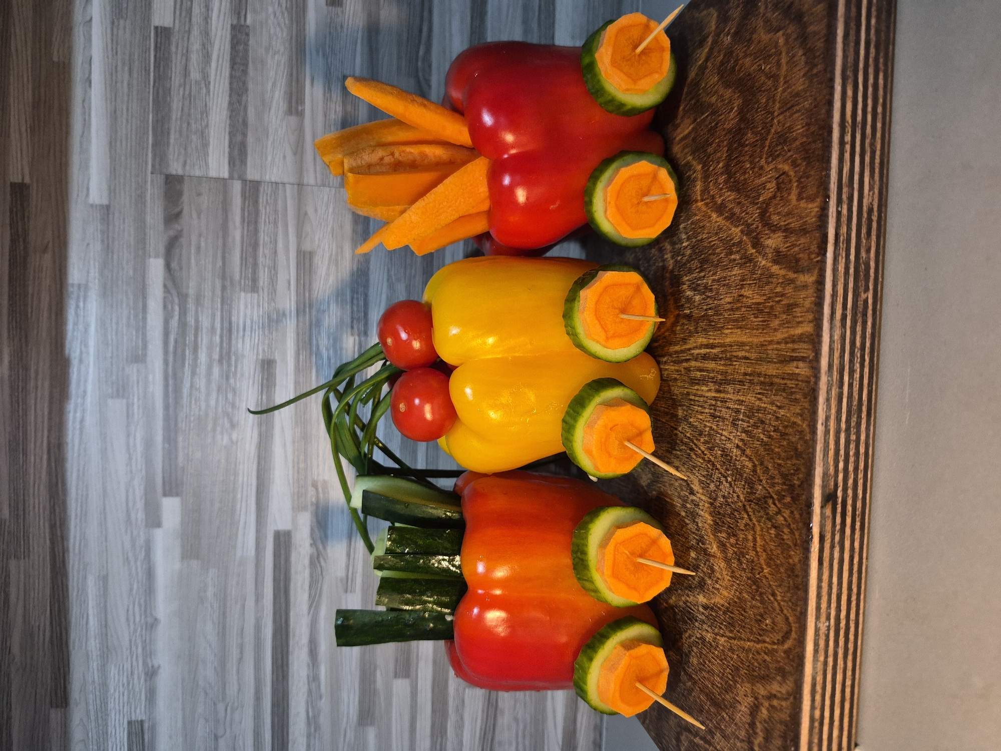 The image shows three bell peppers arranged vertically on a surface with a light gray wood-grain pattern. The top pepper is red and has carrot sticks inserted into it, with cucumber slices containing carrot rounds attached as wheels using toothpicks. The middle pepper is yellow, with cherry tomatoes and green onions inserted, and cucumber-carrot round wheels attached similarly. The bottom pepper is reddish-orange, with cucumber sticks inserted, and cucumber-carrot round wheels. The right side of the image shows a dark brown wooden board.