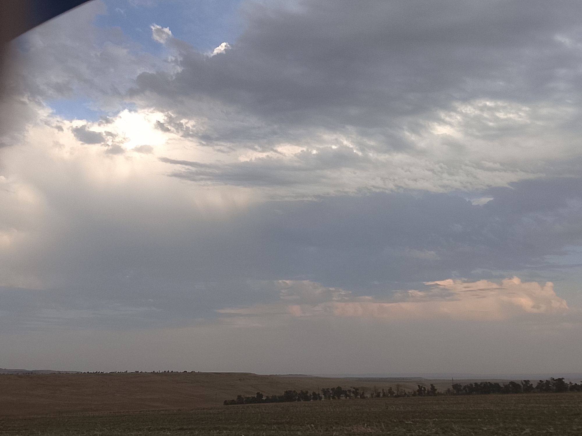 The image depicts a wide, flat landscape under a cloudy sky. In the foreground, a field of dry, brown vegetation stretches across the frame, suggesting it may be late summer or autumn. Further back, a line of scattered trees, appearing dark and silhouetted, runs along the horizon, marking the transition between the field and the distant, hazy land. The sky is dominated by a mixture of gray and white clouds, with a patch of brighter light breaking through the clouds on the left side of the frame, indicating the presence of the sun. The overall impression is one of a rural, open expanse under a dynamic sky. The image does not contain any people or specific identifiable structures.