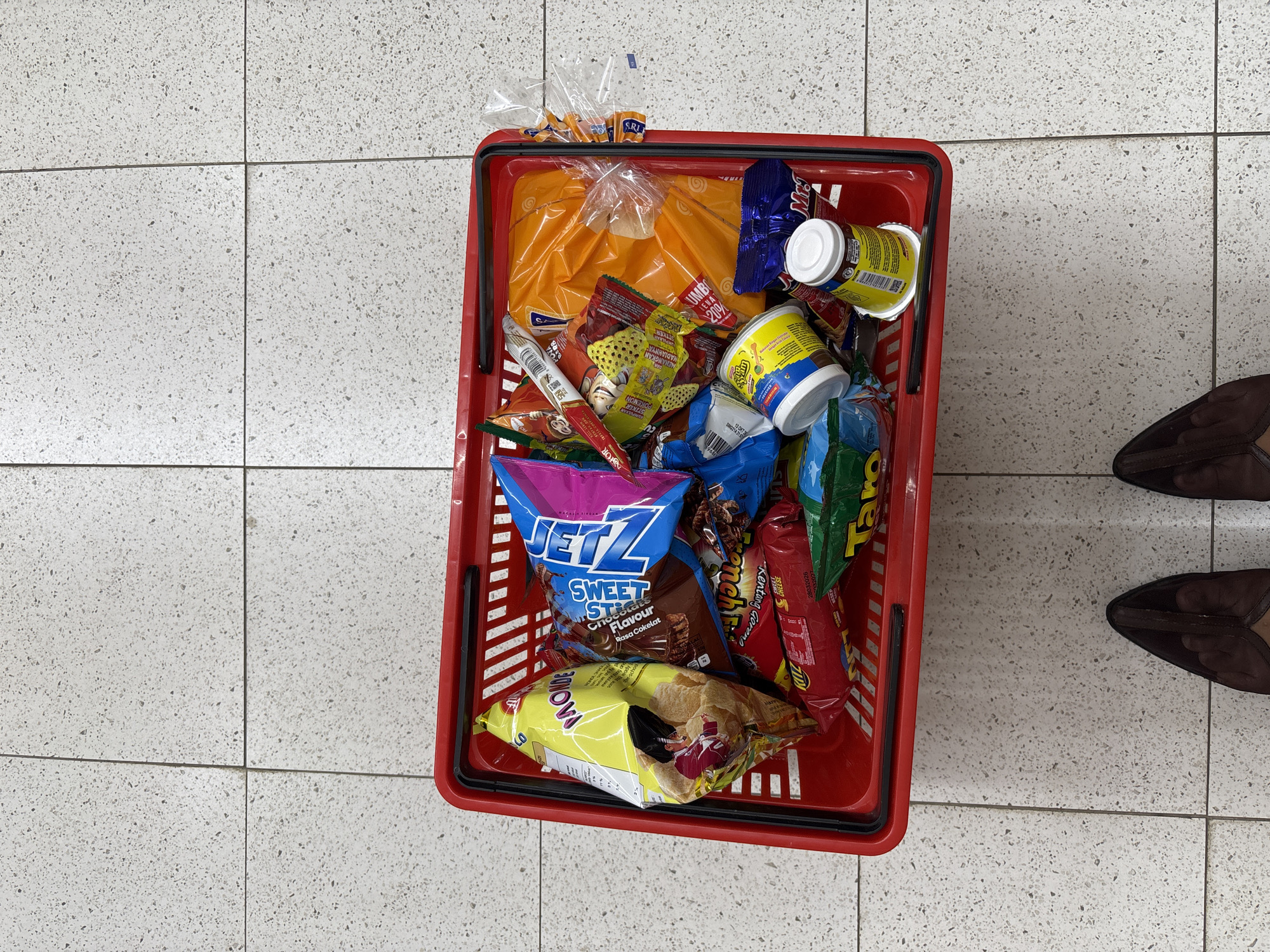 A red shopping basket is on a light gray tile floor, filled with various food items. These include a loaf of bread in an orange plastic bag, multiple cans and packages of snacks, including "JETZ Sweet Sticks Chocolate Flavour," "BONOVI," "Taro" chips, and "Kentang Goreng" chips. The basket is positioned near the edge of the frame, with a person's feet, shod in dark brown flats, partially visible to the right. The scene appears to be within a store in Sukabakti, Indonesia based on the product packaging language.