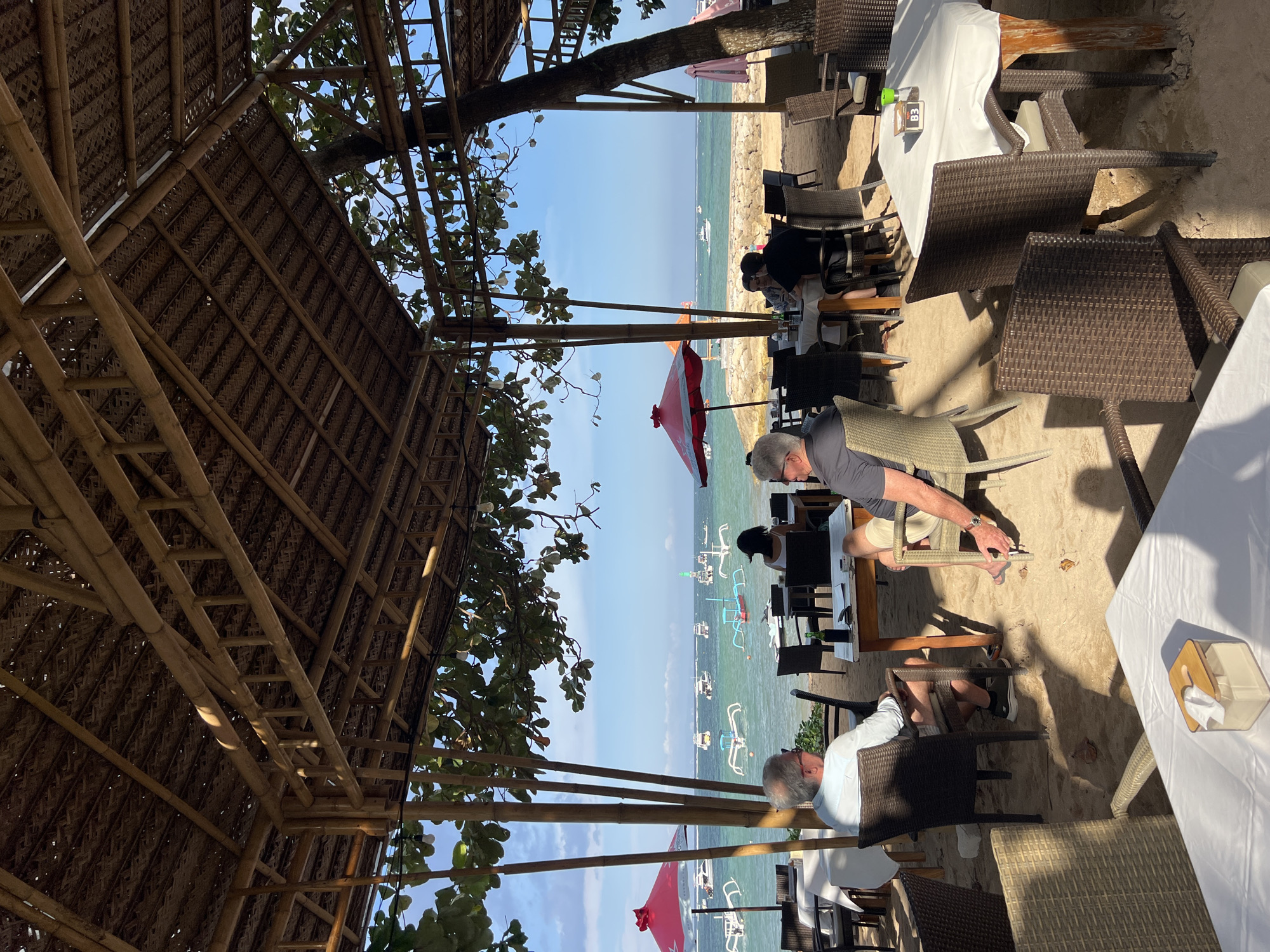 This is a photograph taken at a beachfront restaurant in Denpasar, Indonesia. The scene depicts an outdoor dining area under a thatched roof supported by bamboo poles. In the foreground, tables are set with white tablecloths and chairs, some occupied by diners. A man with gray hair is seated at a table in the left foreground, facing away from the camera. To his right, another man wearing shorts and a grey t-shirt is also seated at a table. In the middle ground, several tables and chairs are visible, with people dining and one woman facing away from the camera. The background shows a calm, turquoise ocean with numerous boats, including traditional Balinese outrigger boats, anchored offshore. The sky is clear and blue. Red and pink umbrellas are dotted around the seating area. The overall atmosphere is relaxed and tropical, typical of a beachside establishment in Denpasar.