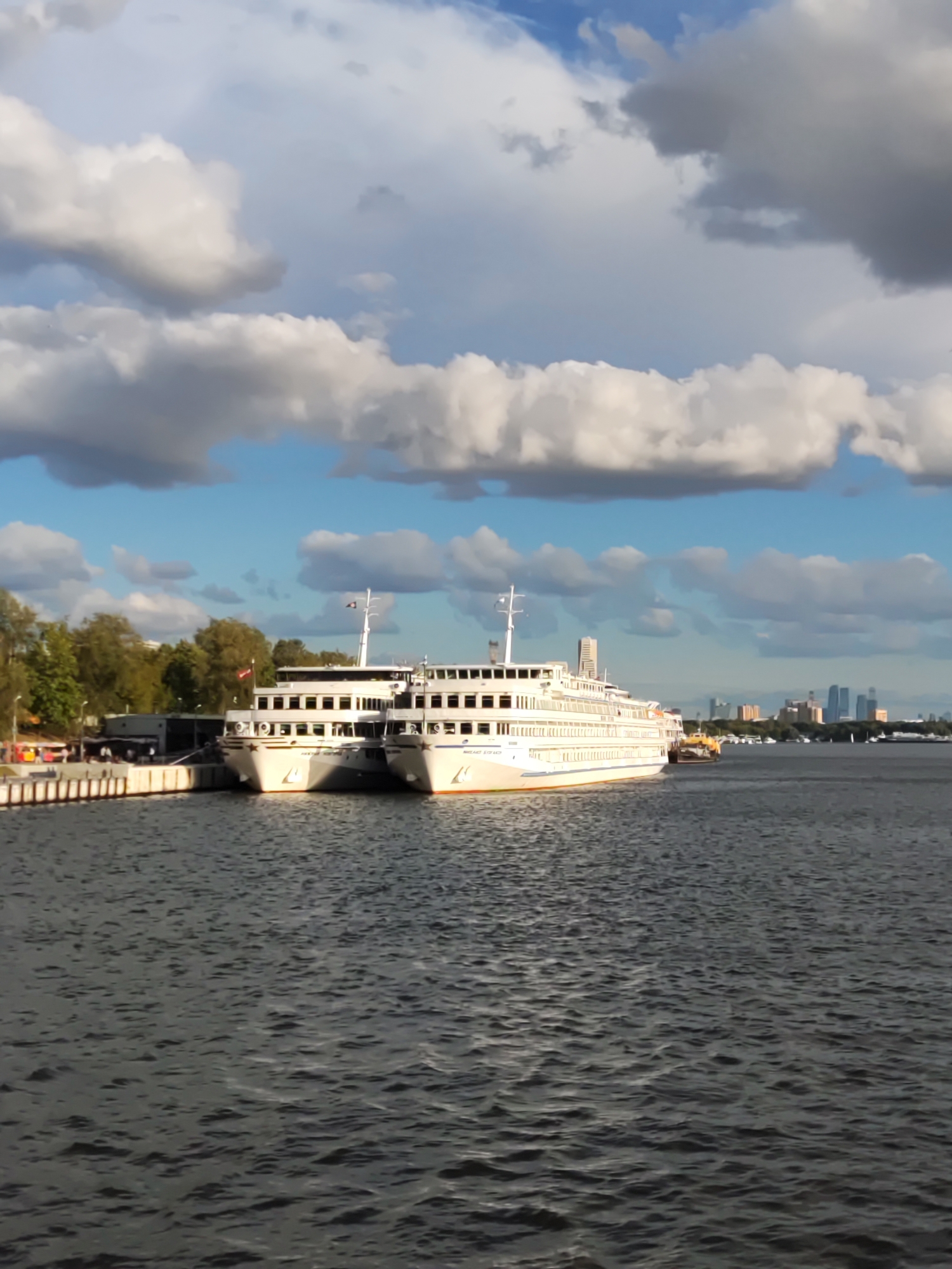 A wide shot shows two white passenger ships docked side-by-side at a pier on a river, with a city skyline in the background, at the Khimki Reservoir on the Moskva River in Khimki, Russia. The ships are long, multi-decked vessels with numerous windows. The closest ship has text on its hull that appears to read "MIKHAIL 1910". The pier is concrete and has some fencing and structures along its edge. Trees are visible on the left side of the pier. The water is choppy and dark. The sky is blue with dramatic, fluffy white and gray clouds. In the distance, a cluster of tall buildings forms a city skyline. The overall impression is a sunny day with a slight breeze.