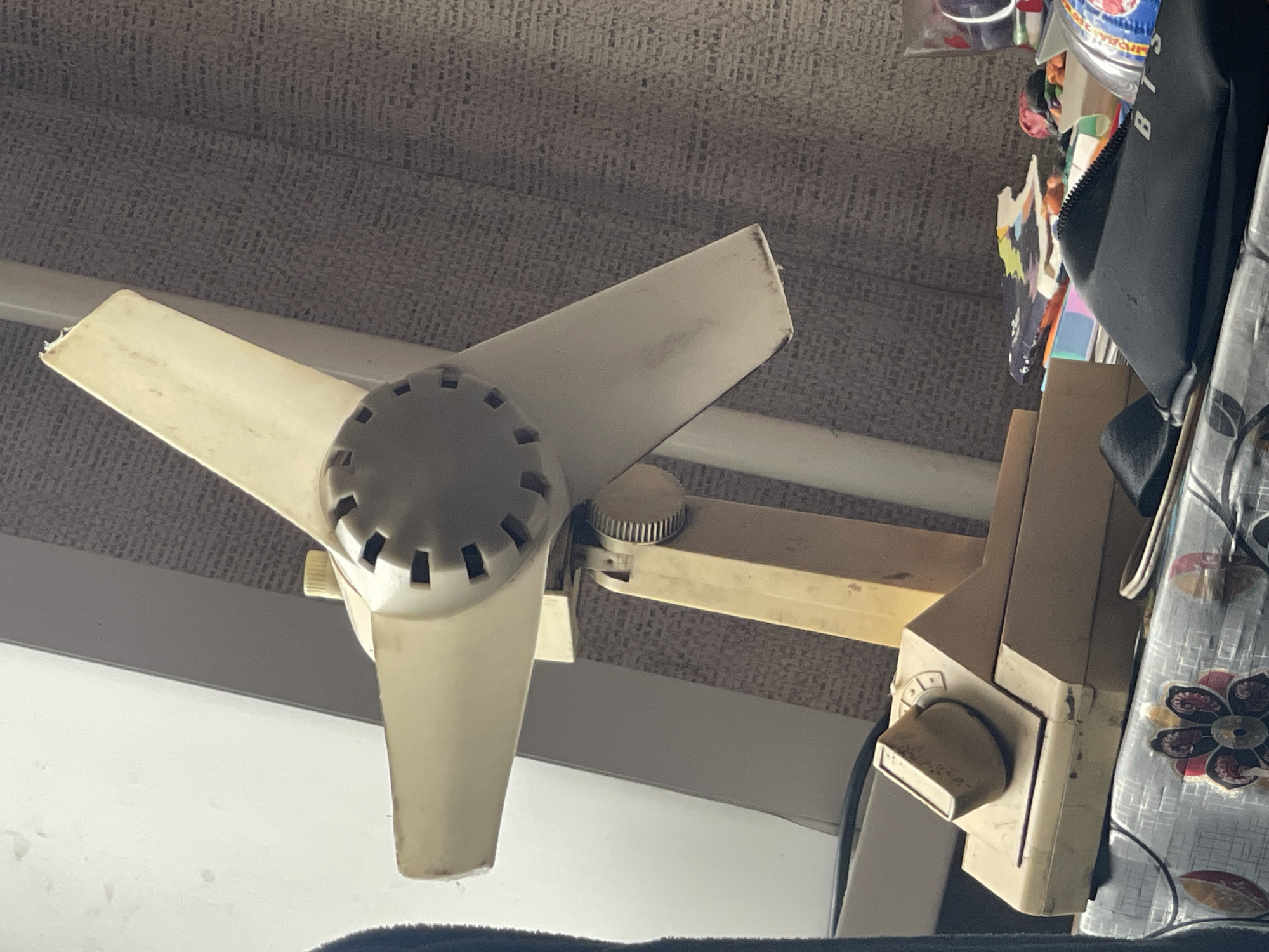 This is a close-up, eye-level, medium shot of a vintage, cream-colored electric fan. The fan is angled, showing its three blades and a round, slotted center. The fan is mounted on a beige, adjustable stand which is attached to a beige, rectangular base with a knob on top. The fan and its base are dusty. The background consists of a light gray wall and a textured, gray curtain. To the right of the fan, on a patterned tablecloth, are several objects including a black pouch with "BTS" written on it, colorful artwork, and a clear plastic container. The scene appears to be indoors, likely in a residential setting in Sibirskiy, Russia.