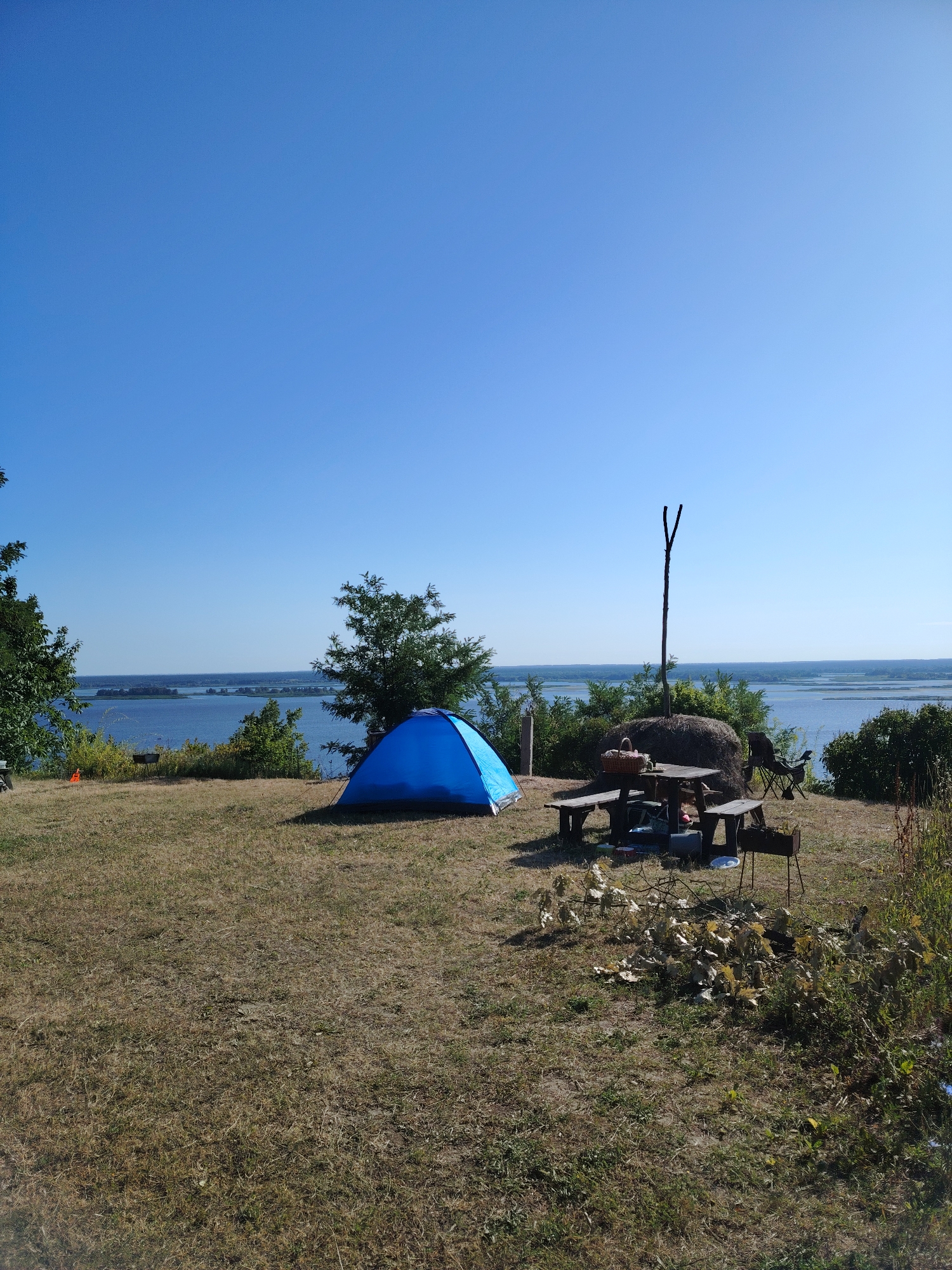 This is a wide-angle, eye-level, outdoor, daytime photograph of a campsite set up on a grassy knoll overlooking a wide body of water, likely the Kaniv Reservoir near Staiky, Ukraine. The sky is a clear, bright blue. In the foreground, dry, matted grass covers the ground. To the left, a bright blue tent is pitched. Further to the right, a wooden picnic table with two benches is set up. On the table, a basket and what appears to be food are visible. Behind the table, a large bale of hay is present, with a wooden stake protruding from its top. A portable grill stands to the right of the picnic table. In the middle ground, lush green bushes and trees line the edge of the knoll. The background features a vast expanse of calm, blue water, with a distant, tree-lined shore visible under the clear sky. No people are present in the scene.