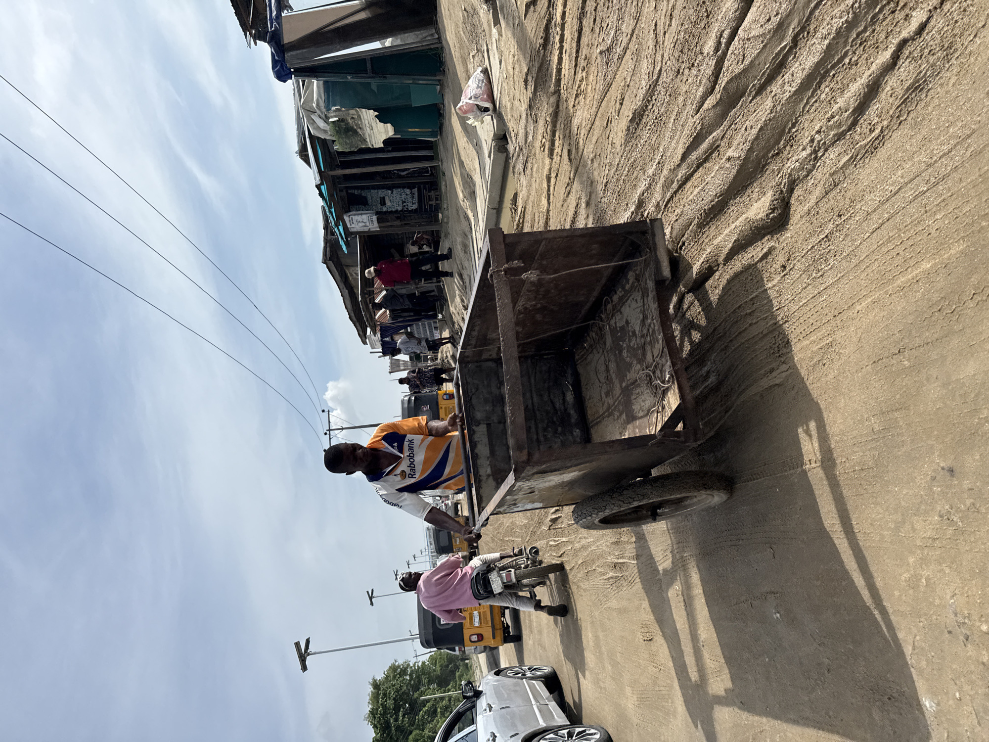 The image shows an outdoor scene in Ado, Nigeria. A man wearing a white t-shirt with orange and blue patterns is standing on the street, pushing a rusty, metal, four-wheeled cart. A man in a pink shirt sits on a motorcycle beside the cart. Other people and several yellow auto-rickshaws are visible in the background along with one white car. Buildings with fabric awnings are visible along the left side of the frame, under power lines that stretch across the sky. The ground is a dirt road.