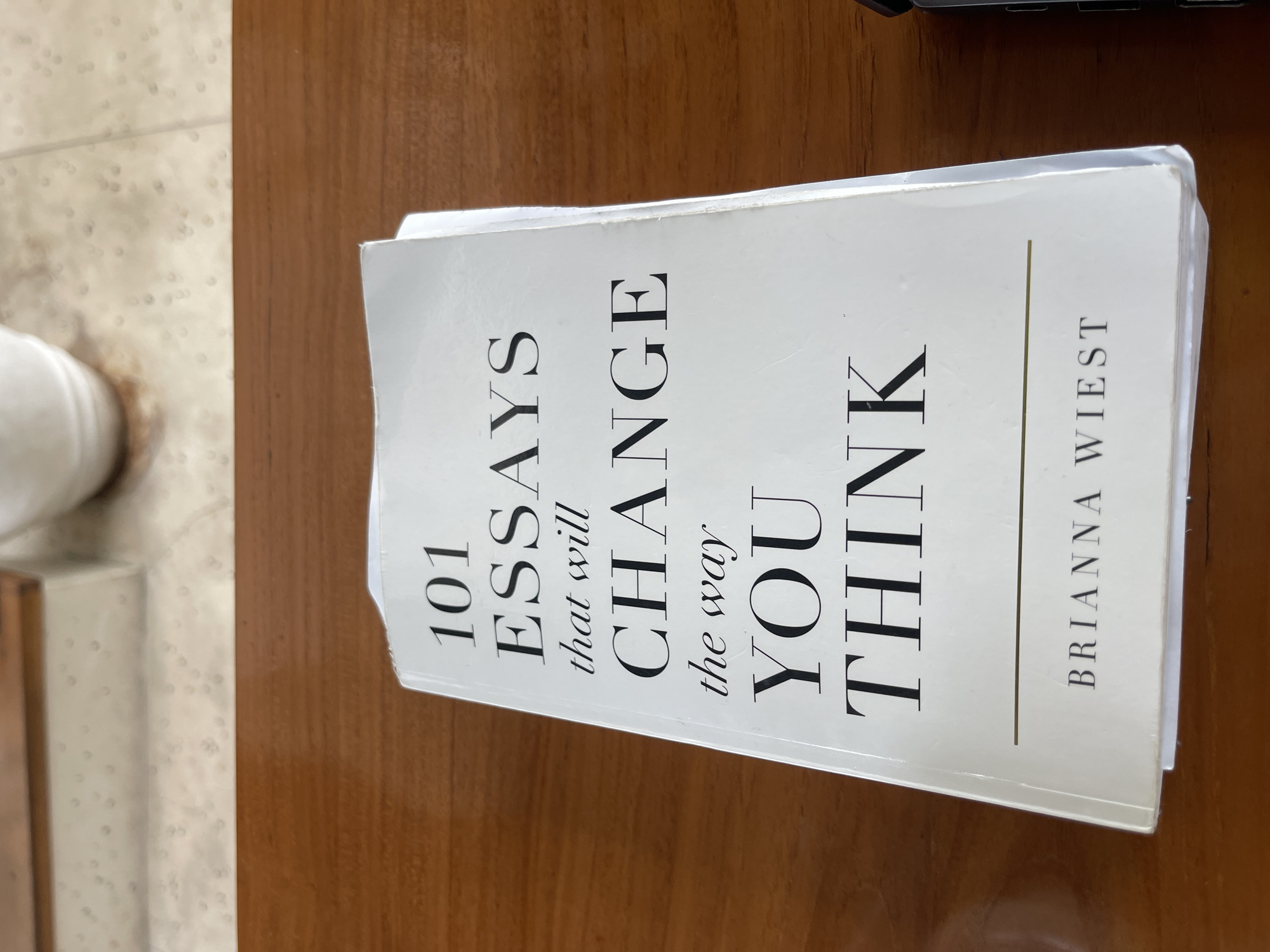 A close-up, eye-level, outdoor shot shows a book titled "101 ESSAYS that will CHANGE the way YOU THINK" by Brianna Wiest. The book is placed on a wooden surface. The cover is white with black text. The title is large and centered on the cover, with smaller text for the author's name at the bottom. The book is slightly angled, with the left side closer to the camera. A thin, horizontal line separates the title from the author's name. The background is blurred and appears to be a patterned surface and a wooden structure. This image was taken in North Kuta, Indonesia.