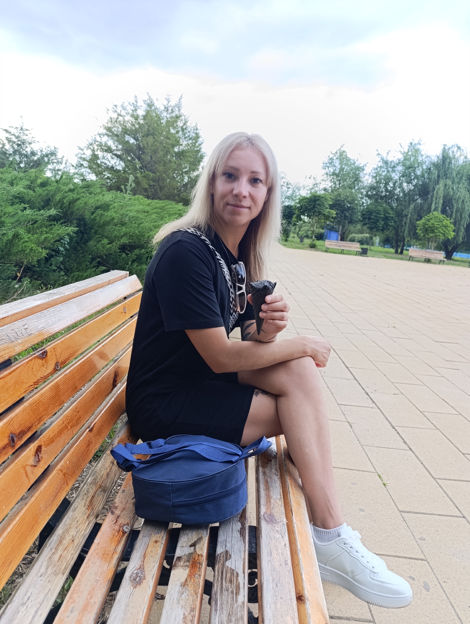 A woman with blonde hair is sitting on a wooden park bench. She is wearing a black t-shirt and skirt, white sneakers, and grey socks. She is holding a black ice cream cone and has sunglasses hanging from her t-shirt. A blue bag is sitting on the bench next to her. The background shows trees, a paved walkway, and a cloudy sky, suggesting it is daytime. The location is Novokuybyshevsk, Russia.