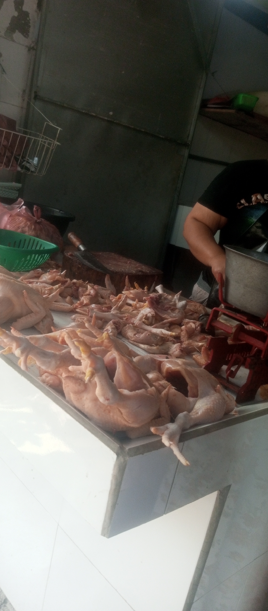 A street-level market stall in Kediri City, Indonesia, displays a large quantity of freshly butchered chickens on a white tiled counter. The chickens, some whole and others appearing to be in parts, are arranged across the surface. Behind the chickens, a large wooden butcher block with a cleaver resting on it is visible, along with a red and silver scale. A person, wearing a black t-shirt, is partially visible to the right, with their arm reaching towards the scale. Various other items, including a green plastic basket, a large red plastic bag, and a metal bucket, are scattered around the stall. The background consists of dark gray walls and shelving, with a metal rack containing a wire basket hanging on the left. The overall scene suggests a typical butcher shop or food market.