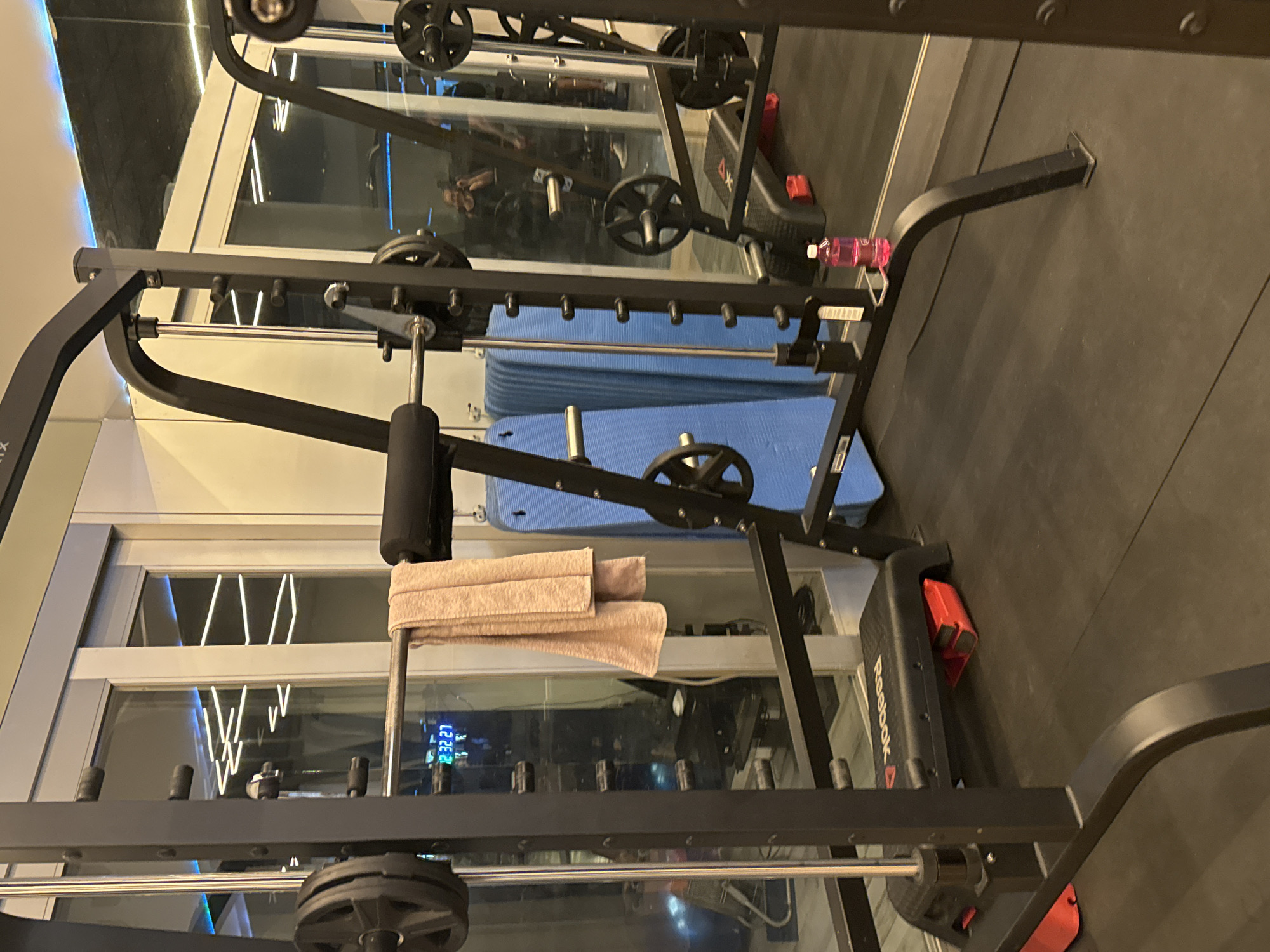 This is an indoor image of a gym with a smith machine in the foreground, a workout bench with a red base, and blue exercise mats stacked behind the smith machine. A pink towel hangs from the smith machine's bar. A bottle of pink liquid is placed on the smith machine's frame. The gym appears to be modern, with glass doors and reflective surfaces in the background, showing lights and possibly other gym equipment. The floor is covered with black gym mats. The image does not provide any information to suggest it is located in Murcia, Spain.