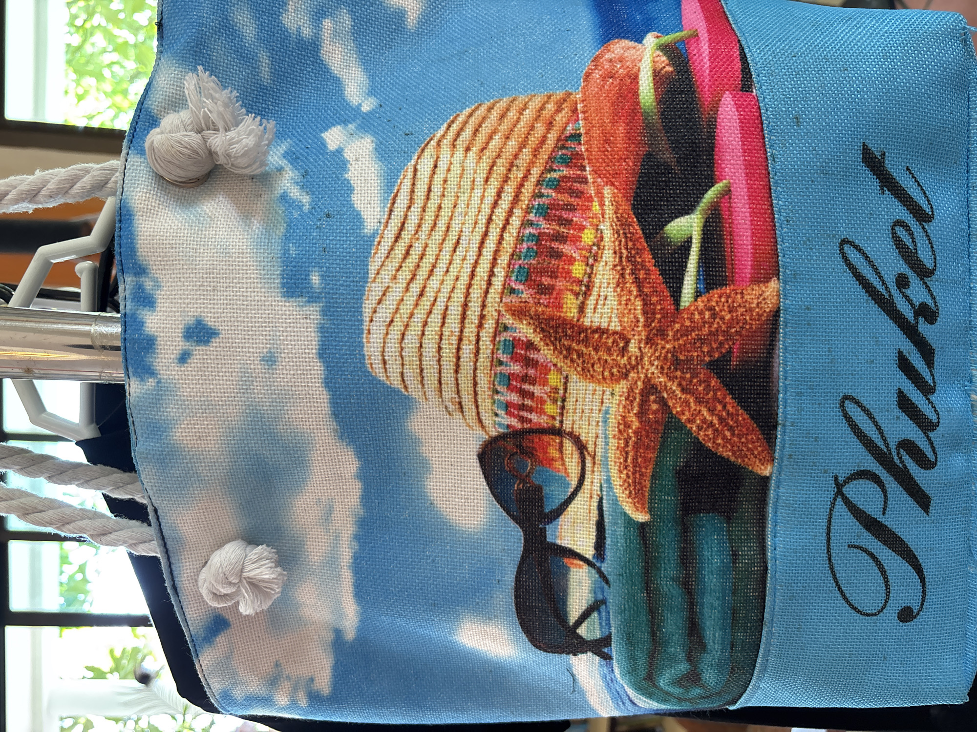 A close-up shot shows the front of a canvas beach bag with a printed tropical scene. The scene features a straw hat, sunglasses, a starfish, flip-flops, and towels against a backdrop of blue sky with white clouds. The word "Phuket" is embroidered in black script on a blue band at the bottom of the bag. The bag has white rope handles. The background is slightly out of focus, showing a window with greenery outside.