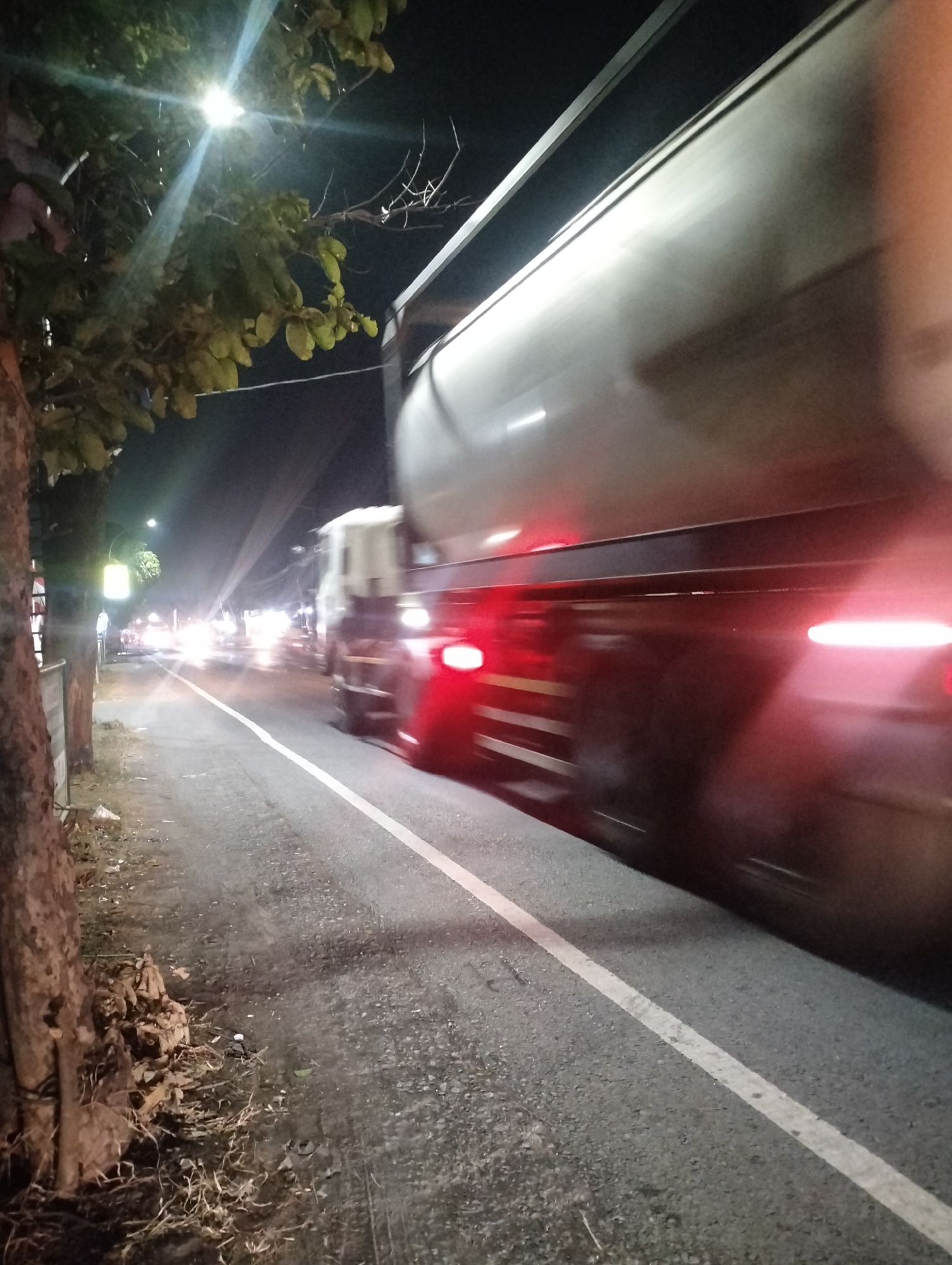 The image captures a nighttime scene on a road in Driyorejo, Indonesia, characterized by motion blur and artificial lighting. The primary subject is a large truck, possibly a tanker, in motion, dominating the right side of the frame. Its side is a dull, metallic gray, with streaks of red light emanating from its rear, suggesting the truck's taillights and possibly reflecting light from other vehicles. The motion blur indicates a significant speed.
The road itself is asphalt, with a clear white lane divider marking the right edge of the truck's path. To the left of the road, a sidewalk or verge is visible, with a large tree trunk in the foreground on the far left, its bark textured and dark. Fallen leaves and debris are scattered around the base of the tree and along the edge of the road. Further down the road, the lights of other vehicles and streetlights create a bokeh effect, with bright, diffused circles of light against the dark night sky. One prominent streetlight with a bright, starburst-like glare is visible above the foliage of the tree in the upper left quadrant. The overall atmosphere is one of a busy nocturnal roadway.