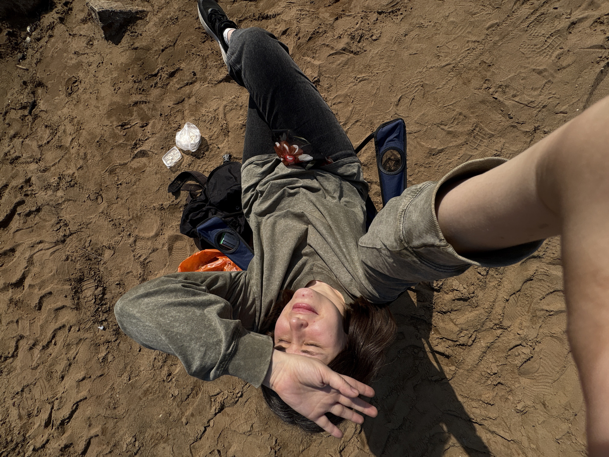 A woman lies on her back in a sand-covered area near the city of Chaikovsky, Russia. She wears jeans, a sweatshirt, and sneakers, and uses her hand to shield her eyes from the sun. A backpack, a partially visible orange bag, a floral-patterned phone case, a blue chair, and discarded plastic packaging are near her. The sand shows impressions from shoes and other disturbances. A hand and arm reach into the frame on the right side. A pile of rocks is visible in the upper left.