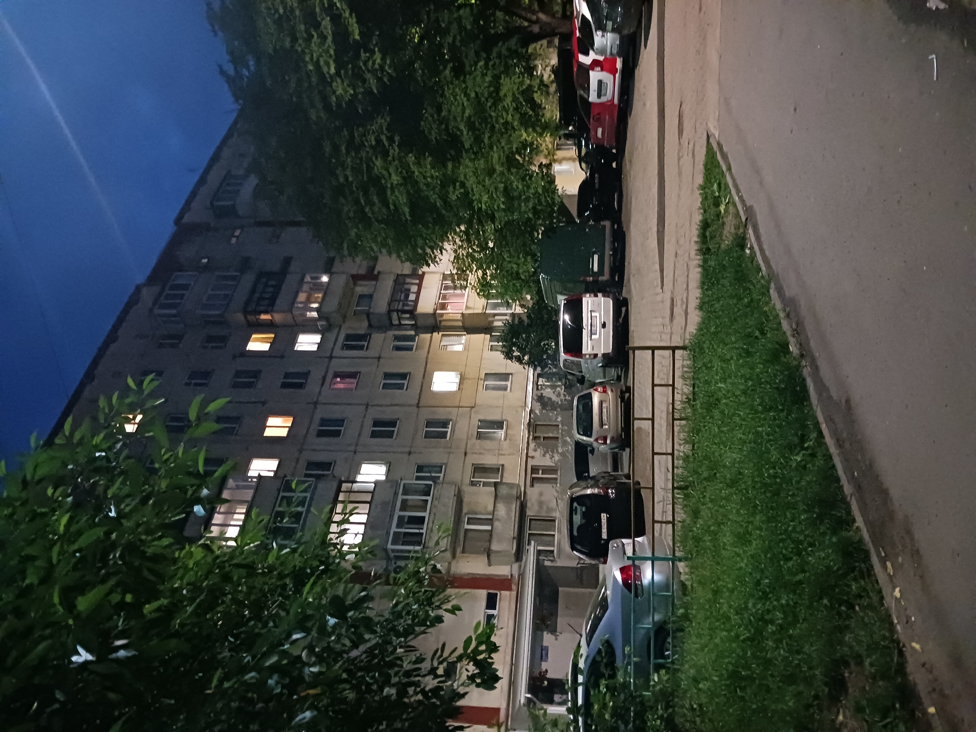 A nighttime exterior scene depicts a multi-story apartment building and a parking lot. The building, a concrete structure with visible balconies, has lights on in several windows, indicating occupancy. In the foreground, a paved area serves as a parking lot, with several cars parked in various positions. Trees and foliage frame the scene, partially obscuring the building and contributing to the overall ambiance. The sky above is dark, with a hint of twilight remaining, and a faint streak, possibly a contrail, is visible. The scene appears to be in a residential area of a city.