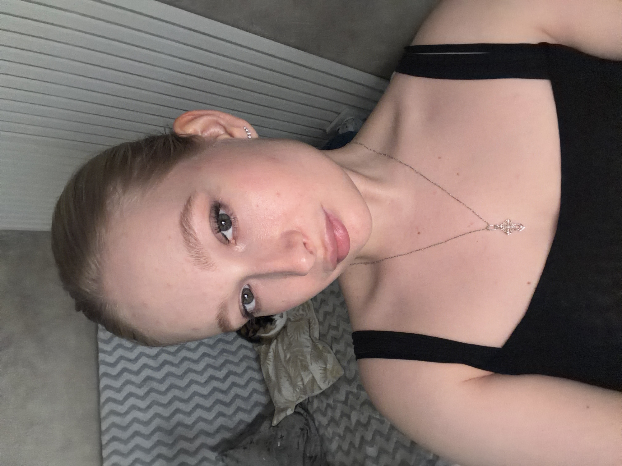 A woman with light skin is facing the camera. She has brown hair and hazel eyes. She is wearing a black tank top and a silver necklace with a cross pendant. She is lying on a grey chevron-patterned blanket with a grey pillow. The background is a white wall with horizontal blinds. The location is Rossiyskiy, Russia.