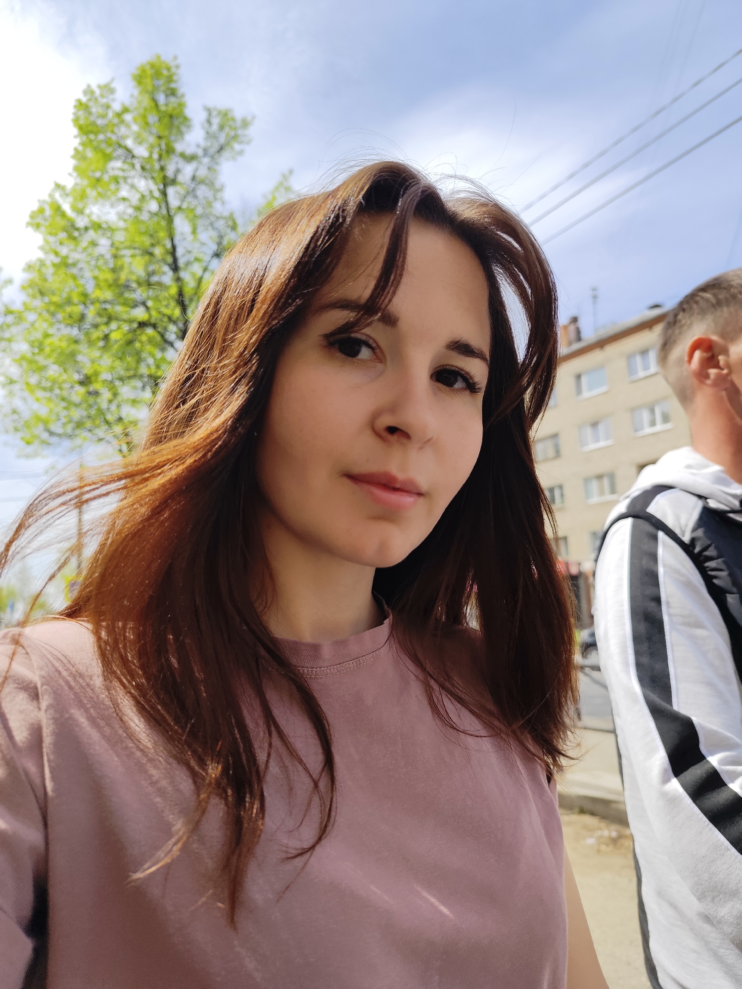 A medium-shot selfie of a young, fair-skinned woman with brown hair. She is wearing a pink t-shirt. A man's head and upper torso are partially visible to the right of her, wearing a white hoodie with a black stripe. In the background, there is a building with multiple windows, a tree with green leaves, and a light blue sky with some clouds. The location is the city of Chaikovsky, Russia. Overhead electric lines are also visible in the background.