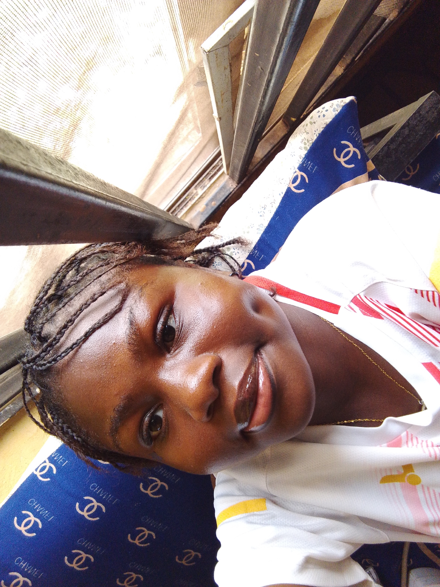 A medium, eye-level shot depicts a young, dark-skinned woman in Nsukka, Nigeria. She's reclined on a blue pillow patterned with repeating stylized logos. She's wearing a light-colored t-shirt with red, white, and yellow accents, and a gold chain is visible at her neck. Her hair is styled into thin braids. The background shows a window with a metal frame and what appears to be a mesh screen. Natural light illuminates the scene.