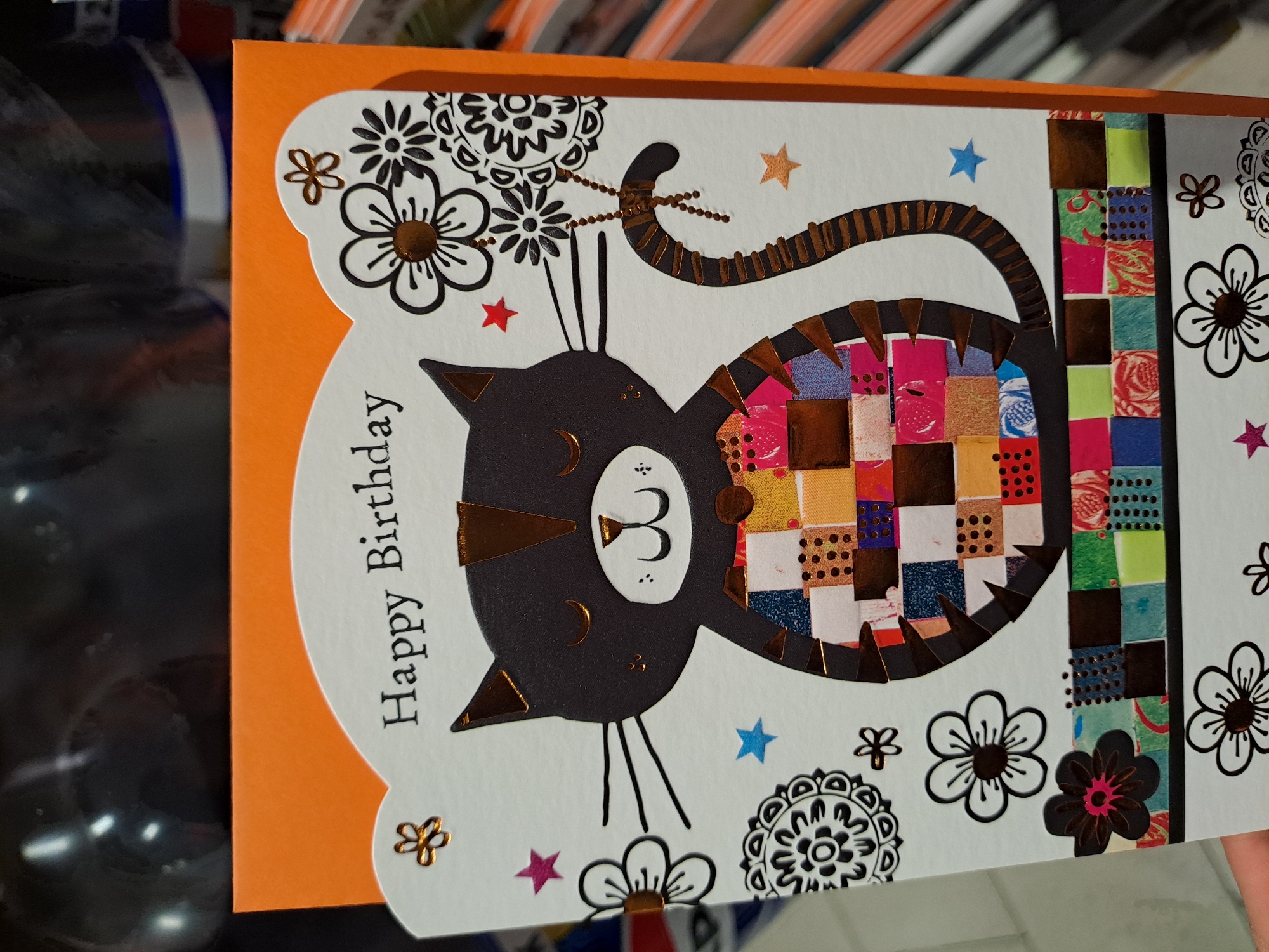 The image shows a "Happy Birthday" greeting card with a black cat design. The cat has gold accents on its ears, nose, and tail. Its body is made up of colorful squares. The card is decorated with floral designs and stars. The background is blurry and appears to be shelves with various items. The card is on an orange surface. There is no indication that this image was taken in Veliko Tarnovo, Bulgaria.