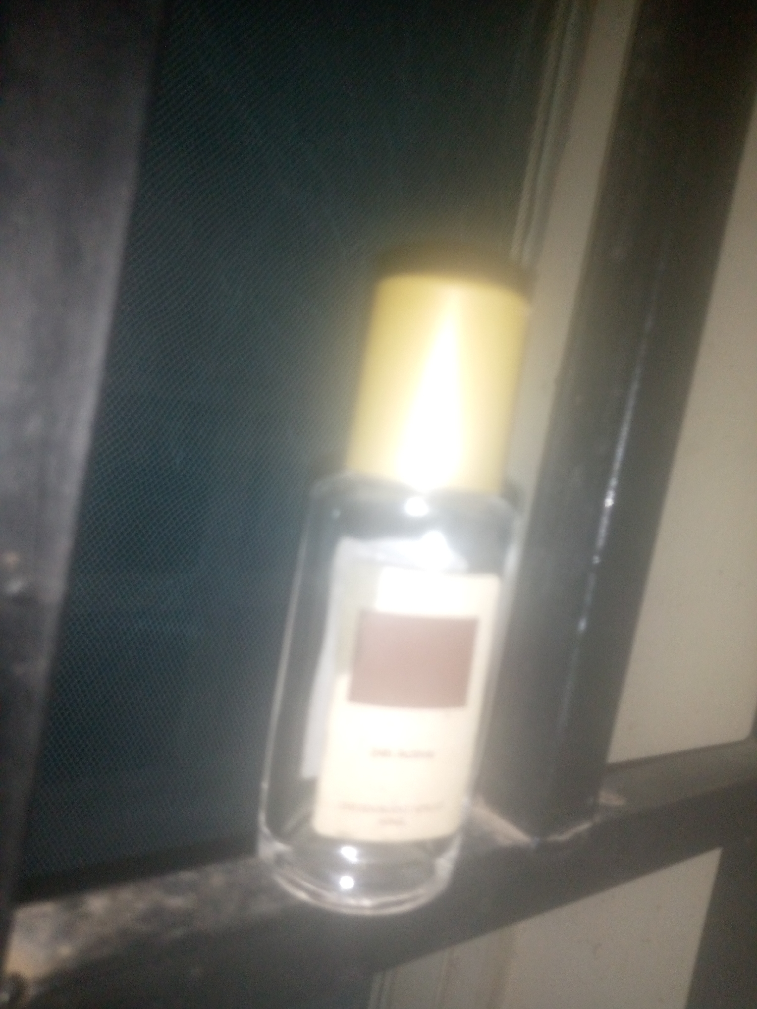 A blurred, dimly lit photograph shows a clear glass perfume bottle with a gold cap resting on a dark, possibly metallic, surface. The bottle has a rectangular label with brown text and a faint white square design. The background is dark and textured, possibly a patterned fabric or screen. The overall impression is of an indoor setting, possibly a shelf or ledge, with the bottle positioned to the right of the frame.