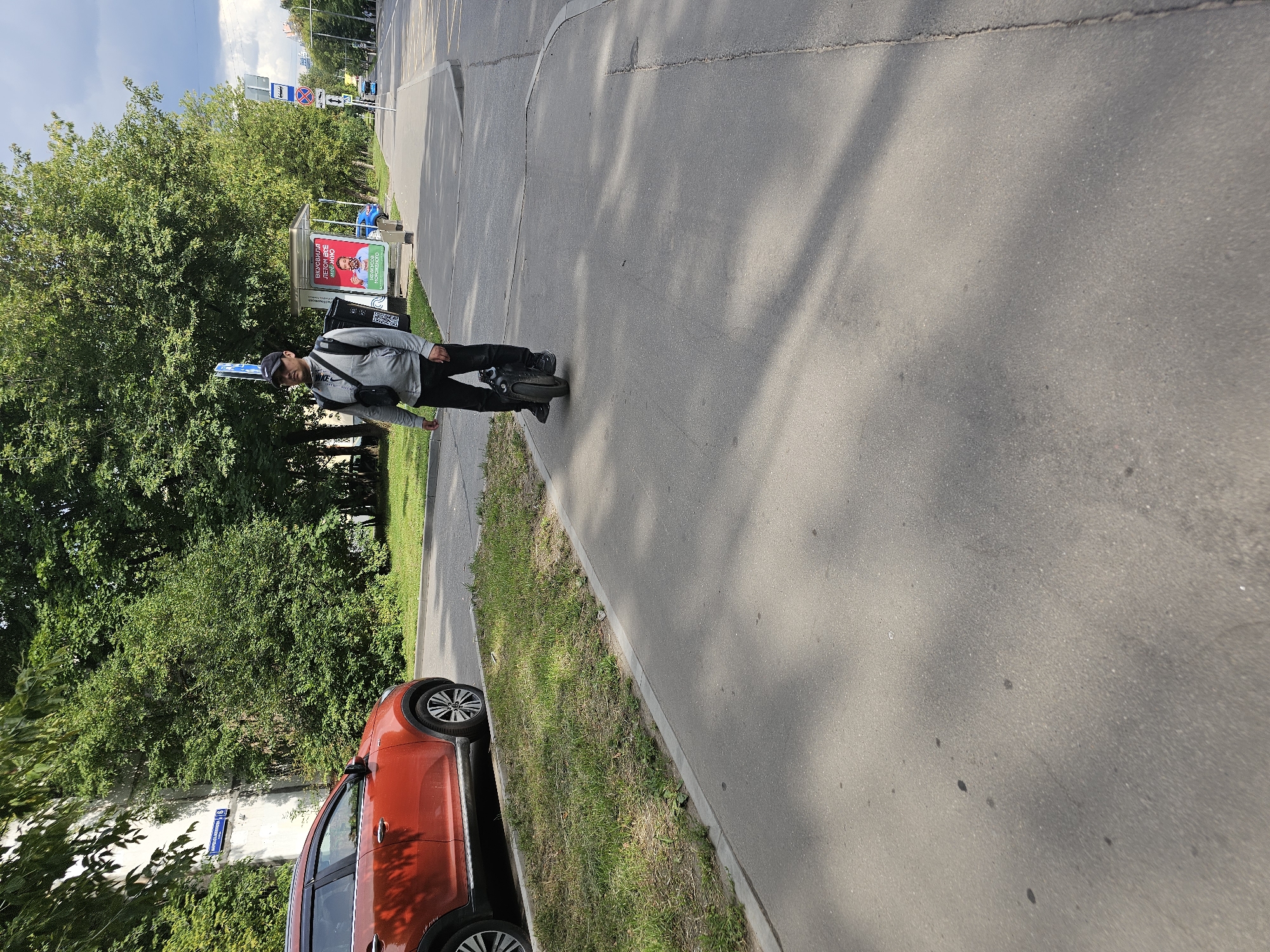 A person is riding a single-wheeled electric unicycle on a paved pedestrian walkway in Khimki, Russia. The person is wearing a grey jacket, black pants, and a black baseball cap. They have a backpack slung over their right shoulder. The walkway is bordered by a strip of grass and then a road. To the left of the walkway, a red SUV is parked. In the background, there are trees, a bus stop shelter with an advertisement, and a street with other vehicles. The sky is cloudy.