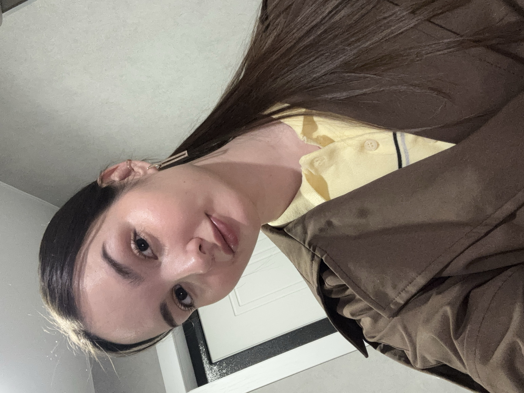 A person with long, dark hair and pale skin is pictured from a low angle. The person is wearing a yellow collared shirt with a black and gray striped trim, and a brown jacket. The background consists of a white wall and a white door with a black frame. There is no indication of the location being Chaikovsky, Russia.