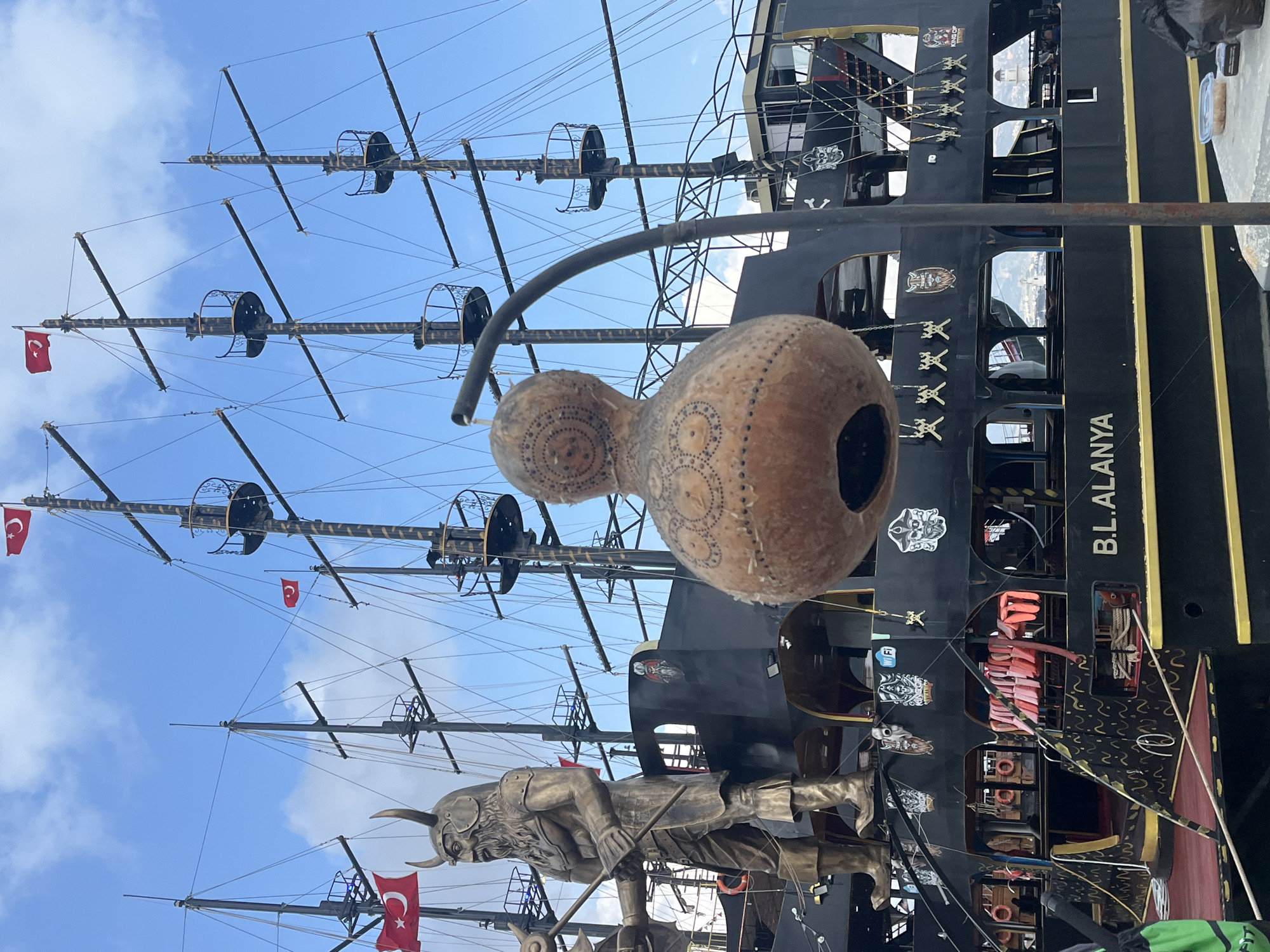 The image shows a detailed view of a tourist pirate ship in Alanya, Turkey. The ship is black with gold trim and prominently displays "B.L. ALANYA" on its side. It features multiple masts with Turkish flags flying from them, along with crow's nests. A bronze-colored Viking statue stands at the ship's bow. In the foreground, a decorative gourd with circular patterns and a large hole is suspended. The backdrop is a blue sky with scattered clouds.