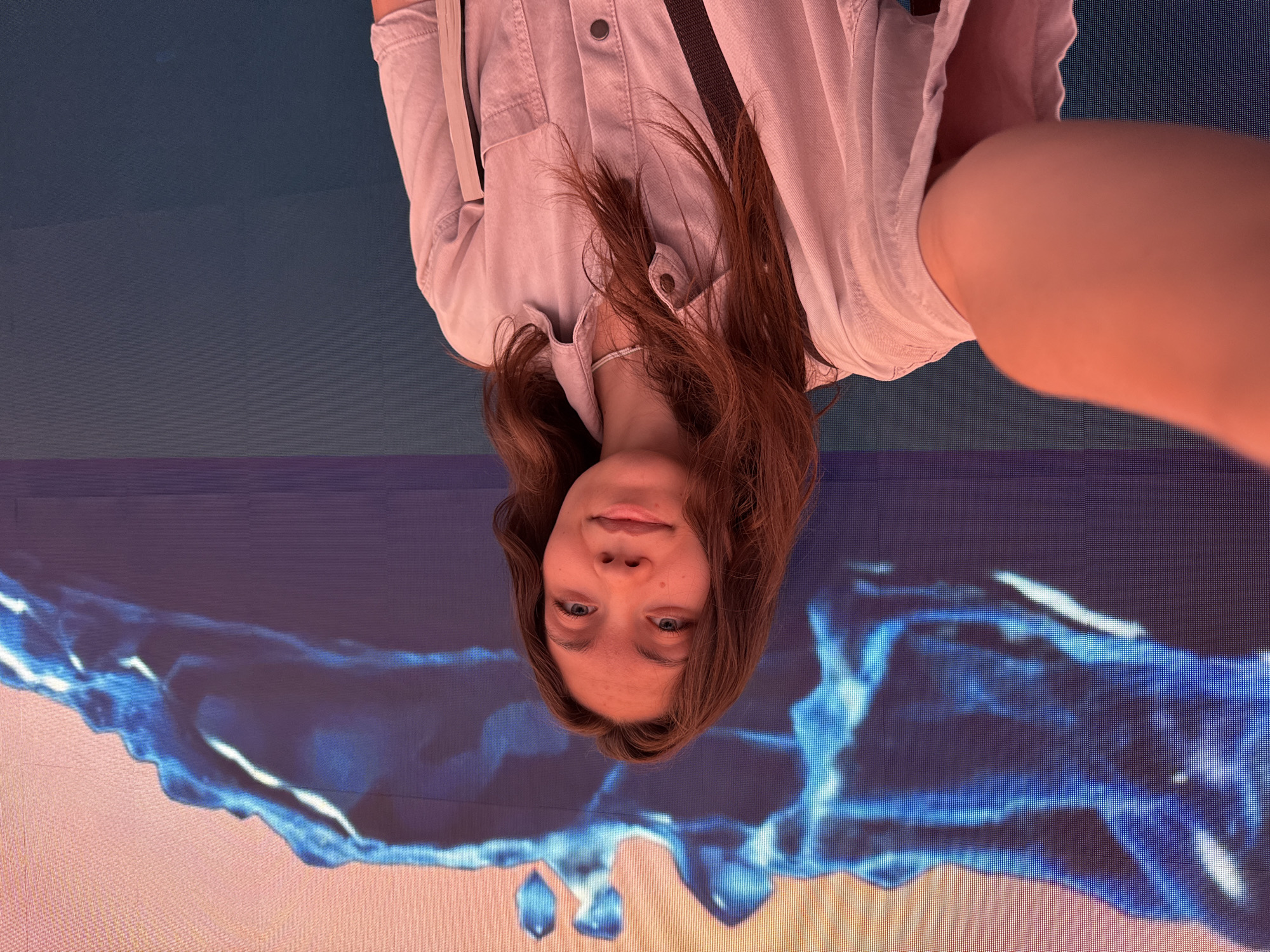 The image shows a young woman with long brown hair and blue eyes, looking directly at the camera. She is wearing a light-colored button-up shirt. The image is taken from a slightly high angle, and the woman's head is upside down. She appears to be standing in front of a large screen displaying a vibrant, abstract blue and orange design that resembles flowing water or smoke. The lighting is soft, creating a warm glow on her skin and hair. There is no discernible text in the image, nor any clear indication of the time of day, weather, or specific location beyond the presence of the screen. The woman's expression is neutral, perhaps a hint of a smile. The overall impression is a candid, somewhat artistic portrait against a dynamic digital backdrop.