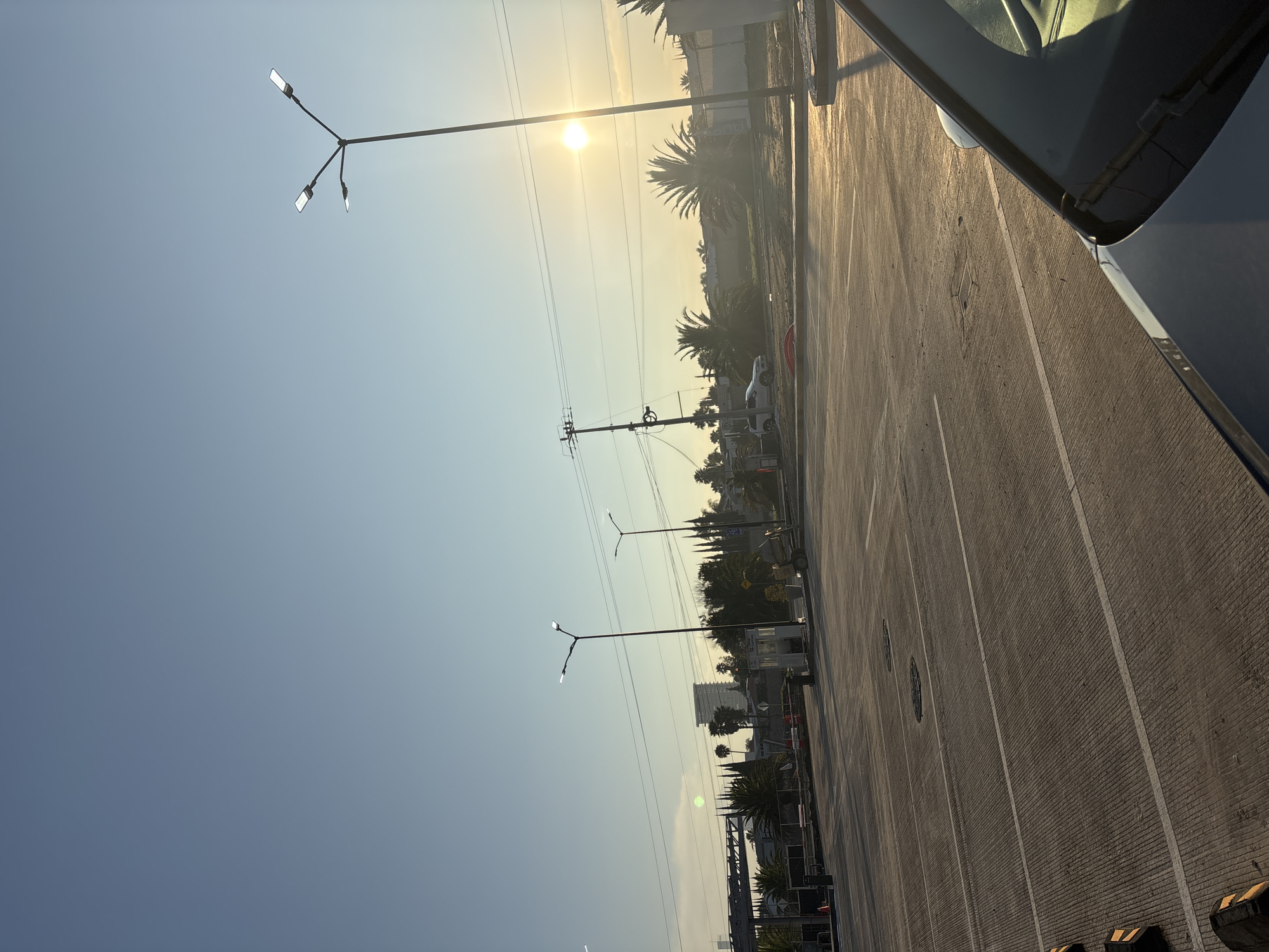 The image depicts an outdoor scene likely in San Bernardino Tlaxcalancingo, Mexico, as the street lighting suggests the region. The foreground shows a portion of a parked vehicle, possibly a car, taking up the right side of the frame. Adjacent to the vehicle is a wide, mostly empty parking lot or paved area with distinct lines marking parking spaces.
The background includes a mix of urban and natural elements. Palm trees are visible, along with buildings and what appears to be a power line stretching across the frame. The sky is a pale blue, with the sun obscured by haze, suggesting either early morning or late afternoon. Several modern streetlights are prominently featured, their poles reaching up into the sky. The overall atmosphere is bright, but hazy.