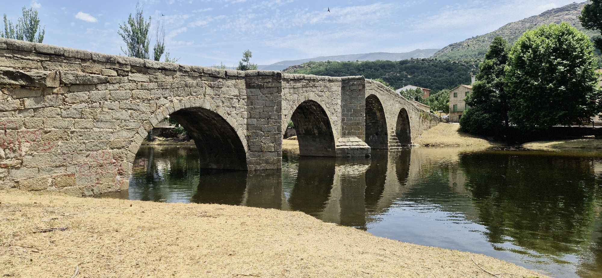A stone bridge with five arches spans a calm body of water, reflecting its structure. The bridge is constructed of rough-hewn, light-colored stones, some of which bear faint red markings. The water's surface is slightly rippled, disturbed by a gentle breeze, and reflects the bridge, trees, and a hazy blue sky dotted with scattered white clouds. The foreground features a dry, sandy bank covered with sparse, golden-brown grass. In the background, beyond the bridge, a lush green hillside rises, topped with more distant, muted mountain ranges. On the right side of the bridge, a two-story building with light-colored walls and green shutters is visible amidst dense foliage and trees. A large, verdant tree stands prominently to the right of the building. The scene suggests a rural or semi-rural setting, possibly in a Mediterranean climate, given the vegetation and the dry ground. The overall lighting indicates it is daytime, with the sun casting shadows. There are no visible people or animals in the immediate scene, though a small bird is captured in flight against the sky.