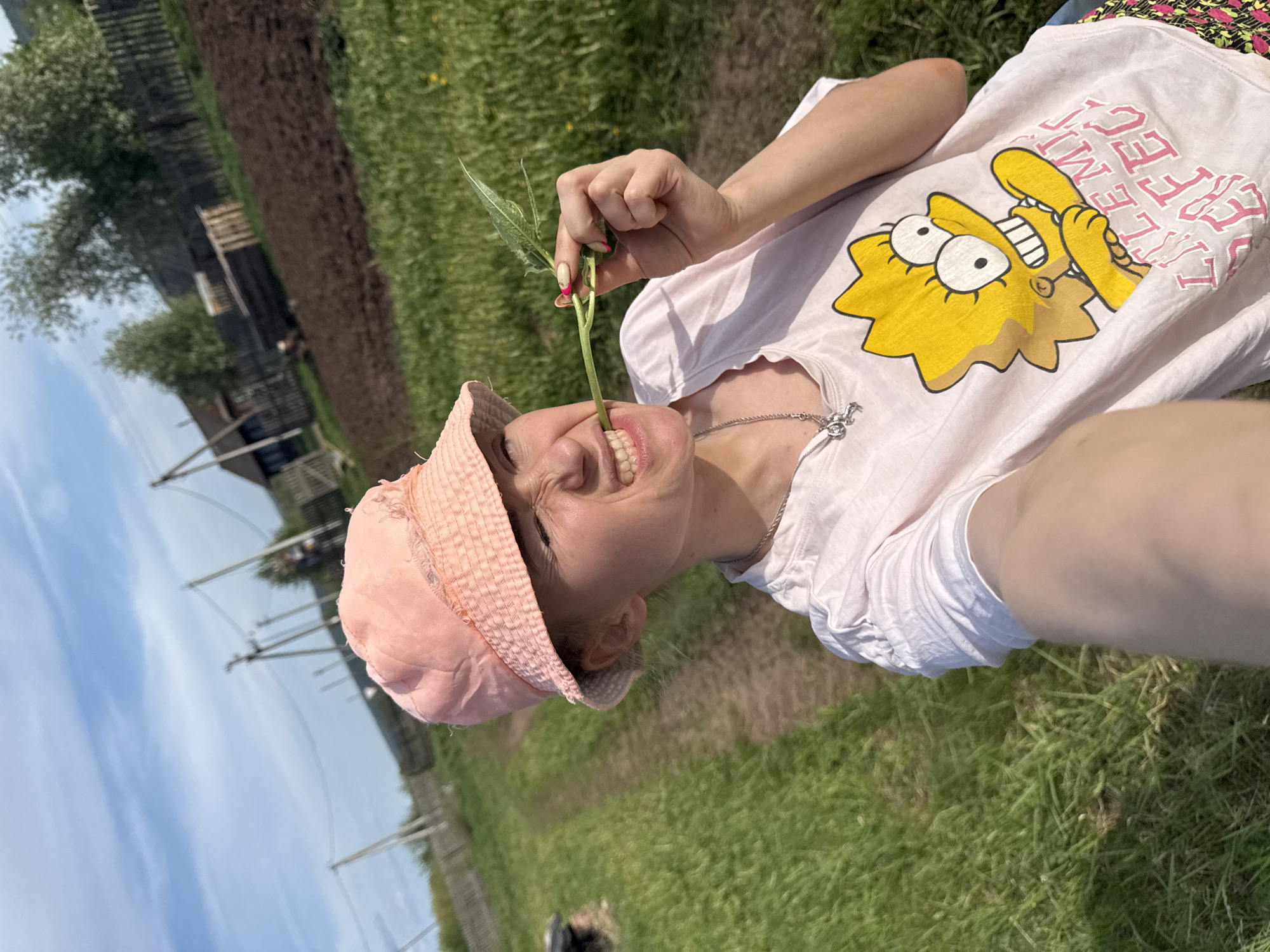 A young woman wearing a pink bucket hat and a white t-shirt with a Lisa Simpson graphic is holding a green plant stem in her mouth. She is outside in a grassy area. In the background, there is a fence, telephone poles, and buildings. The location is Nizhnyaya Chermoda, Russia.
