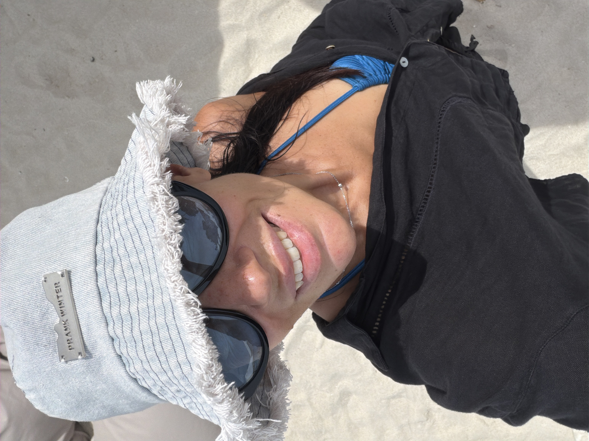 A high-angle, close-up selfie shows a woman from the chest up, wearing a light blue denim bucket hat with frayed edges and a metal plate that reads "PRANK WINTER". She also wears dark sunglasses, a black open shirt, and a blue bikini top. The woman is smiling and her hair is dark and appears wet. The background is a sandy beach, consistent with a location in Baan Chaweng, Thailand.