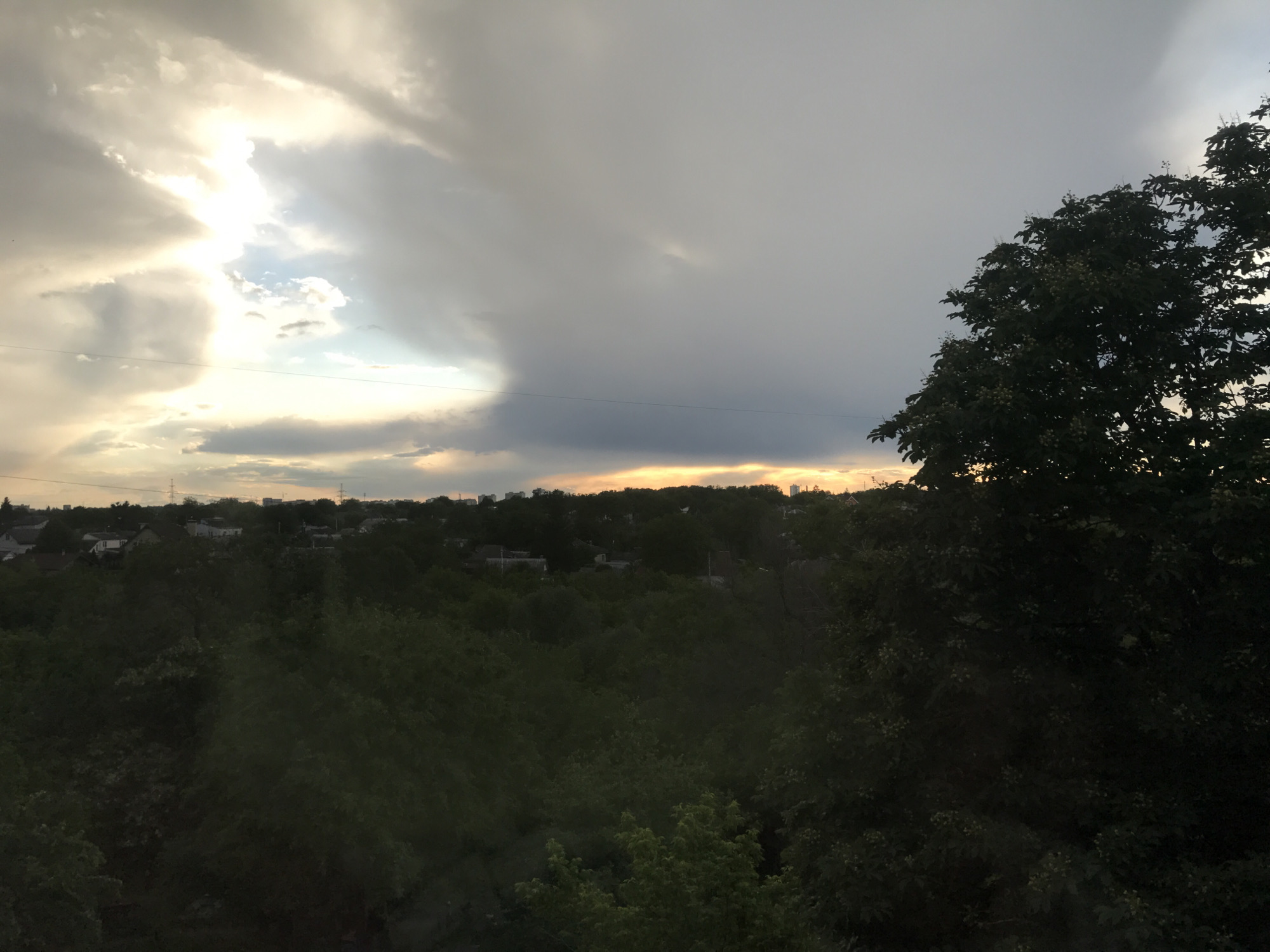 The image depicts an overcast sky over Bezliudivka, Ukraine. A break in the clouds reveals a section of lighter sky and the setting sun. A line of trees separates the foreground and the background. In the background, silhouettes of houses and apartment buildings are visible against the golden horizon. A tall tree occupies the right portion of the foreground.