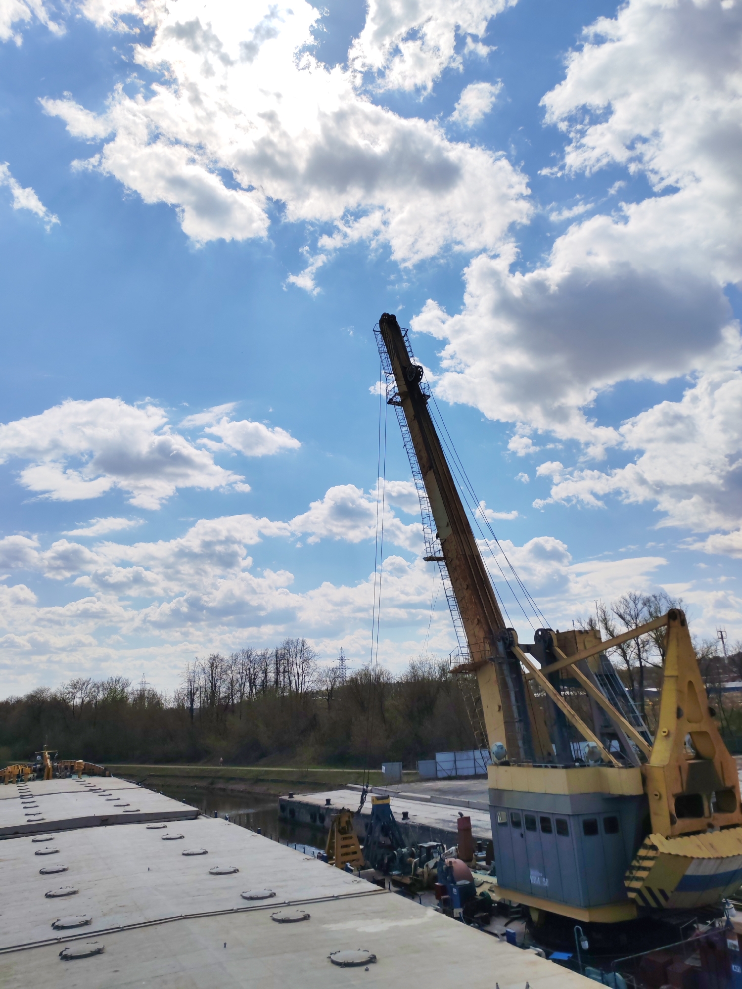 The image shows a crane on a barge in Yakhroma, Russia. The crane, predominantly yellow and gray, is the focal point, extending upwards against a backdrop of a blue sky filled with puffy white clouds. The barge, composed of flat, gray sections, suggests it's being used for transporting goods. In the distance, there are trees, utility poles, and buildings, indicating an urban or industrial area. The water reflects the sky, separating the barge from the distant shoreline.