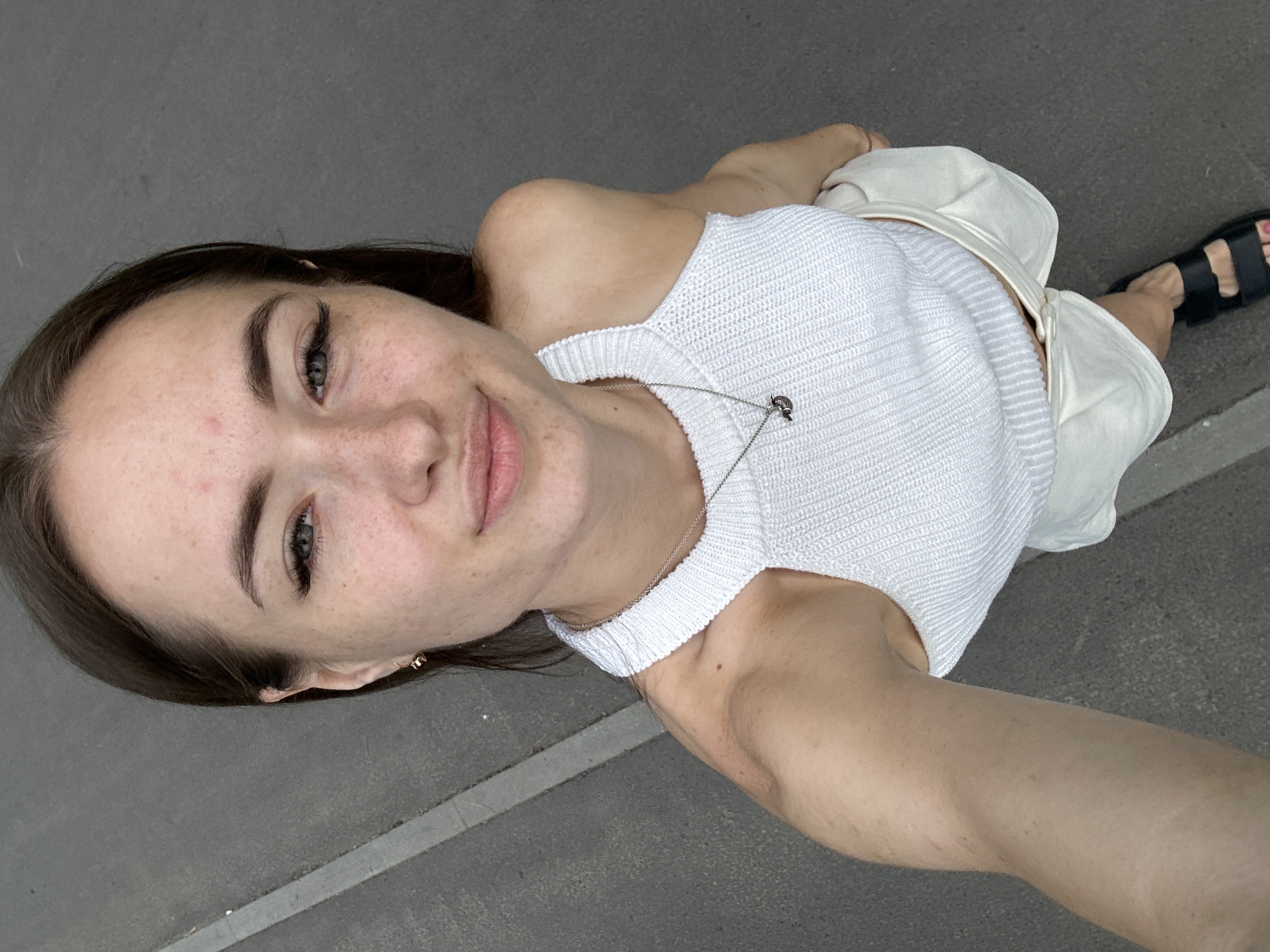 A young Caucasian woman with brown hair and light skin is standing outdoors and taking a selfie. She is wearing a white knit halter top and white shorts. She has a silver necklace with a pendant. She is wearing black sandals. The background is a gray asphalt surface with a light gray stripe. The woman appears to be looking at the camera with a slight smile.