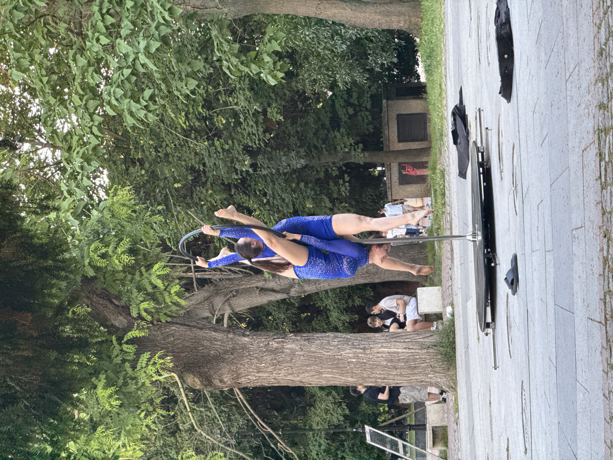 This image captures an outdoor aerial hoop performance in a public park or square in Varna, Bulgaria. Two female performers, both dressed in matching sparkly royal blue long-sleeved bodysuits, execute an aerial hoop routine on a freestanding, portable pole. One performer is inverted, suspended by her knees and hands from the black hoop, while the second performer is positioned above her, also gripping the hoop and extending her legs upward. Both are barefoot.
The performance occurs on a paved surface comprised of alternating light grey and darker grey rectangular stone tiles. A pair of black flip-flops and two collapsed black fabric bags or garments lie on the ground near the base of the portable pole.
The background is densely vegetated with large, mature deciduous trees exhibiting full green foliage. To the left of the scene, behind a substantial tree trunk, a male onlooker in a black t-shirt and light patterned shorts stands adjacent to a white A-frame easel. Further right, two young men are seated on a light-colored concrete bench, observing the performance. One wears a light t-shirt and white shorts, while the other is in a dark jacket. A red fire hydrant is partially visible behind a tree trunk in the mid-ground right. Additional blurred figures are present deeper in the background, including an individual walking in light shorts and a child. The scene is illuminated by natural daylight.