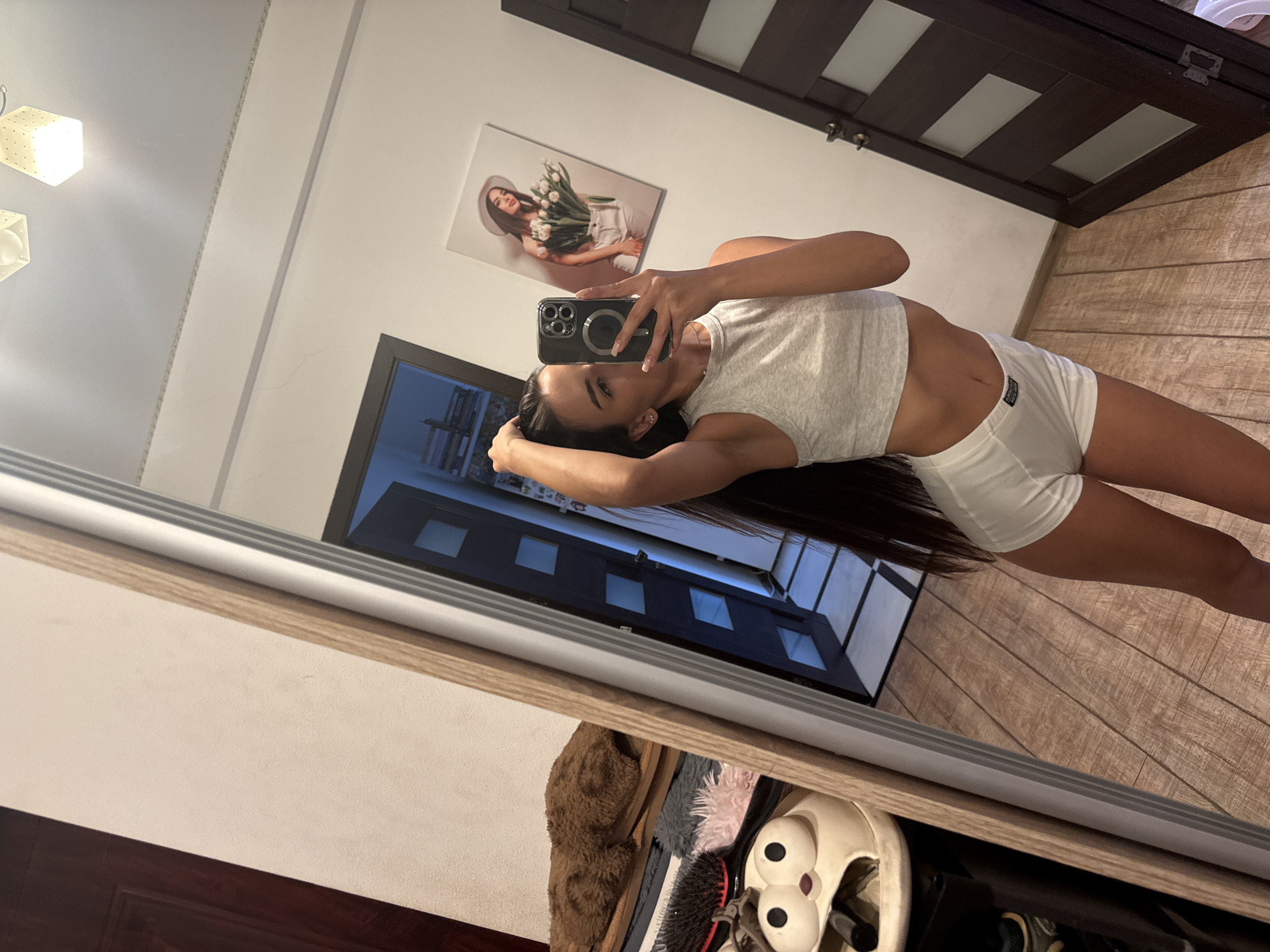 A young woman is taking a mirror selfie in what appears to be a home interior. She is wearing a grey crop top and white shorts. Her hair is long and dark, and she is holding her hair up with one hand. In the background, a painting of a woman with flowers hangs on the wall. To the left of the woman, a collection of items is visible, including what appear to be slippers and hairbrushes. To the right, a dark wooden door with frosted glass panels is seen. The floor is made of wooden planks. The overall impression is that of a casual, domestic setting. There is no specific information in the image to suggest the location is in the city of Chișinău, Moldova.