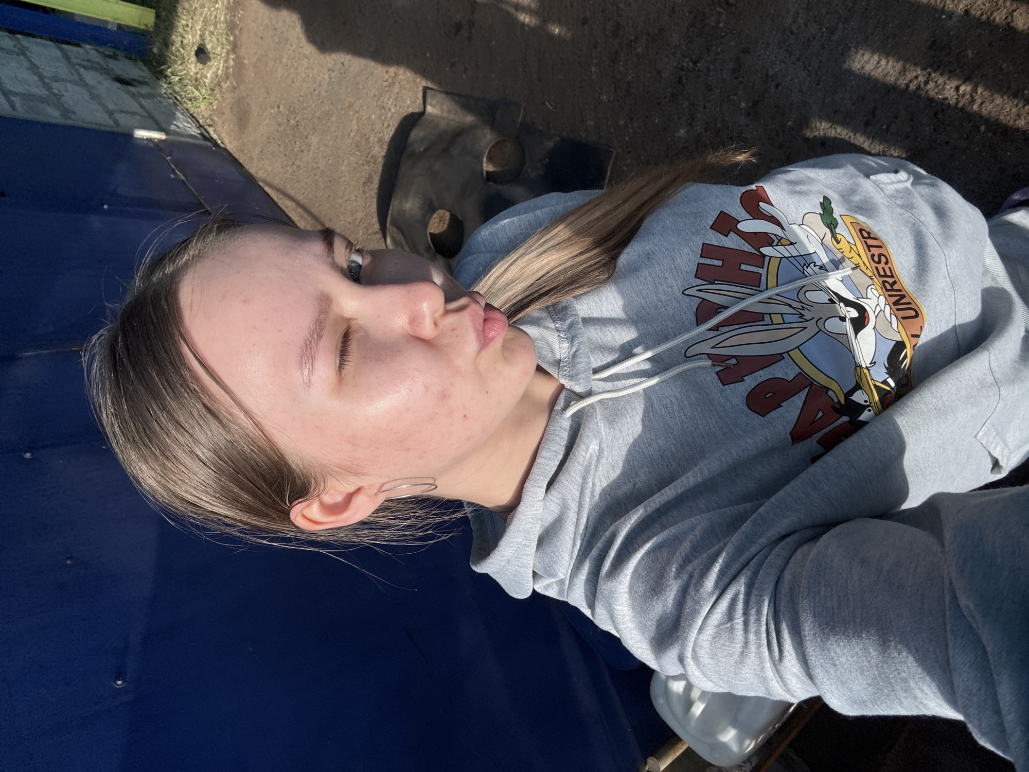 An outdoor, slightly overhead selfie shot captures a young person, likely female, lying down on their side in Zhigalki, Russia, during daytime. The individual has light brown hair that extends over their shoulder and down their back. They are wearing a light grey hooded sweatshirt featuring a colorful graphic. The graphic includes cartoon characters resembling Bugs Bunny, holding a carrot, with partially visible text that reads "TH..." and "UNRESTR".
The person's face is turned towards the camera with a playful expression, eyes open, and lips pursed in a "kissy face" or pout. A simple, thin metallic hoop earring is visible in their left ear. The background shows a mixture of surfaces: a large, dark blue, smooth area that the person's upper body rests on, and beyond that, rough, earthy ground with some patches of grass and a dark, irregularly shaped object.
The scene is brightly lit by strong sunlight, casting prominent shadows, indicating a clear, sunny day. The overall atmosphere is casual and relaxed.