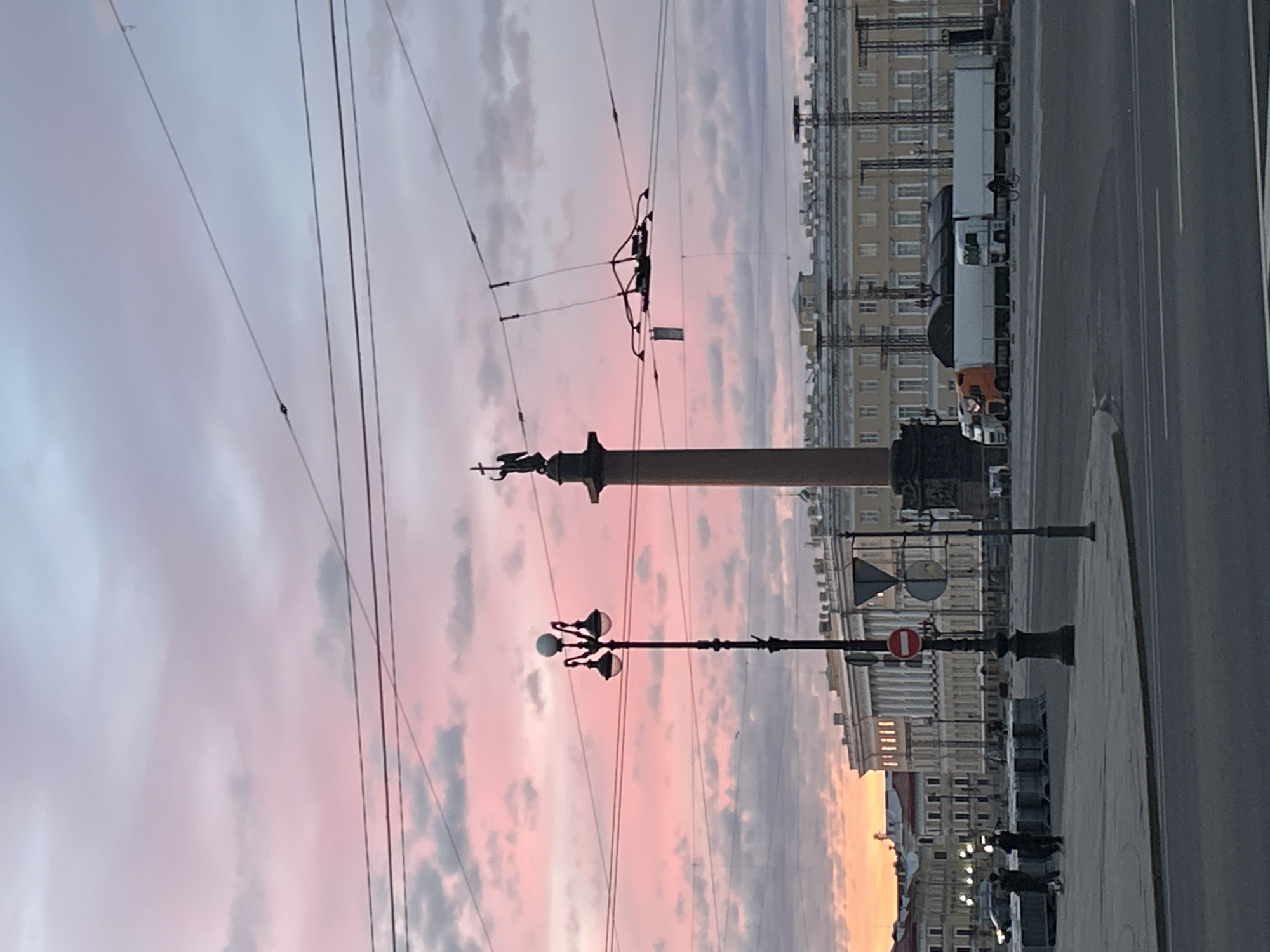 The image depicts a wide shot of a city square at dusk, with a prominent Alexander Column dominating the center. The sky is painted with vibrant pink and purple hues from the setting sun, interspersed with soft clouds. In the foreground, a black lamppost with multiple globes stands tall. The background features a grand, light-colored building with classical architecture, possibly a government or museum building, with numerous windows and ornate details. Several trucks are parked in a row in front of the building. On the left side of the image, two individuals are visible walking on a sidewalk. Overhead, intricate network of wires, likely for transportation or communication, crisscross the sky. The overall scene suggests a public space in an urban environment during twilight hours.